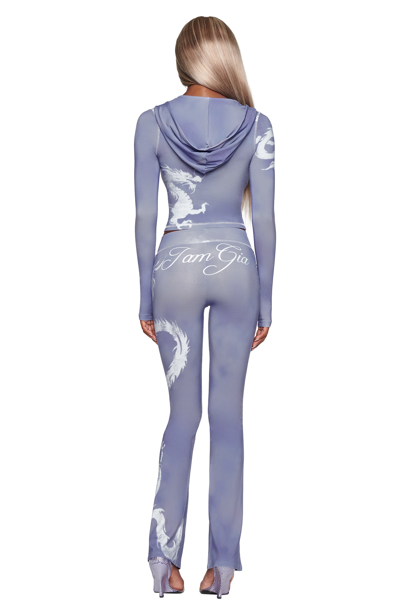 A woman with long, straight blonde hair stands with her back to the camera, wearing a fitted, sheer purple jumpsuit featuring bold white dragon motifs and “I AM GIA” text on the back. Her striking look channels the distinctive style of I.AM.GIA’s BLARE TRACKPANT in Steel Blue Dragon, known for its unique dragon design and signature attitude.