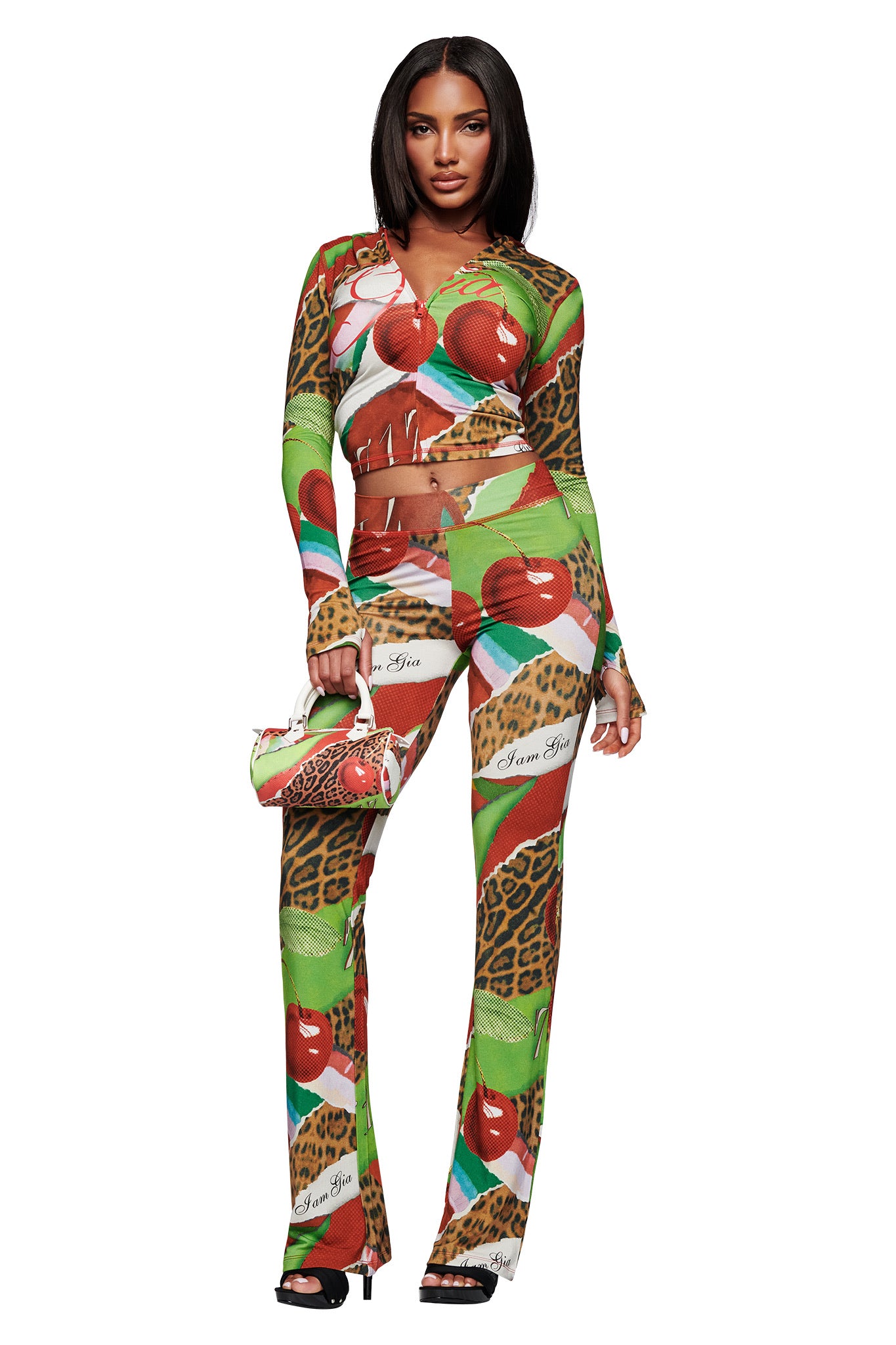 A woman faces forward in the I.AM.GIA BLARE HOODIE - WILD CHERRY, a colorful long-sleeve crop hoodie with an apple and leopard print. She carries a matching bag and pairs the look with black high-heeled sandals.