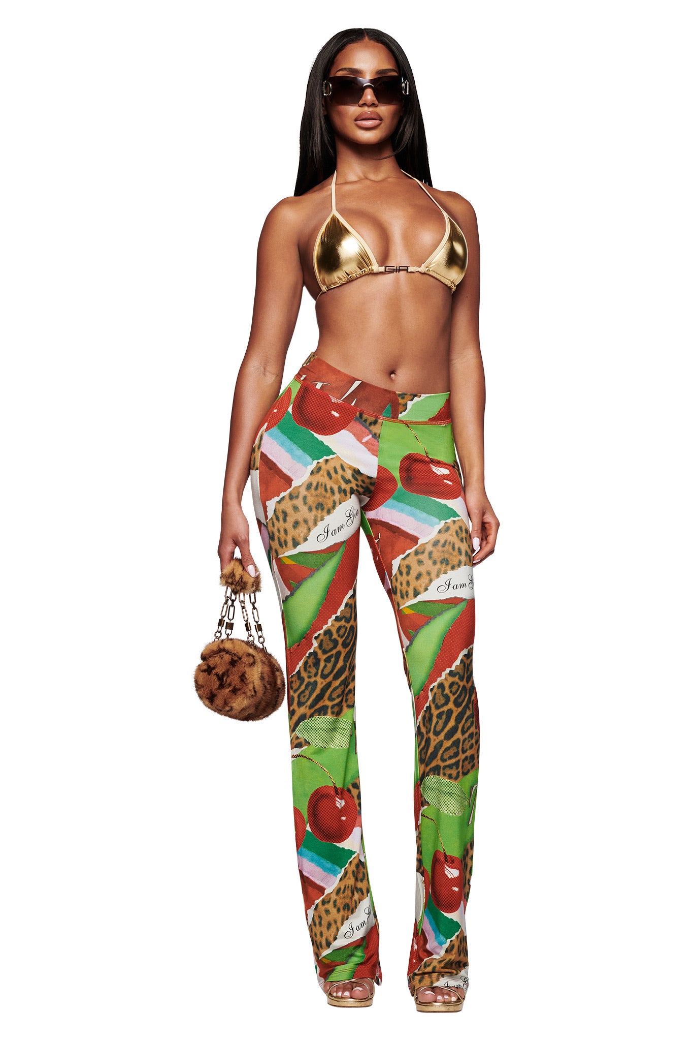 A woman poses in a metallic gold bikini top, I.AM.GIA BLARE TRACKPANT - WILD CHERRY with animal motifs and bamboo fabrication, clear heels, sunglasses, and a small furry handbag against a white background.