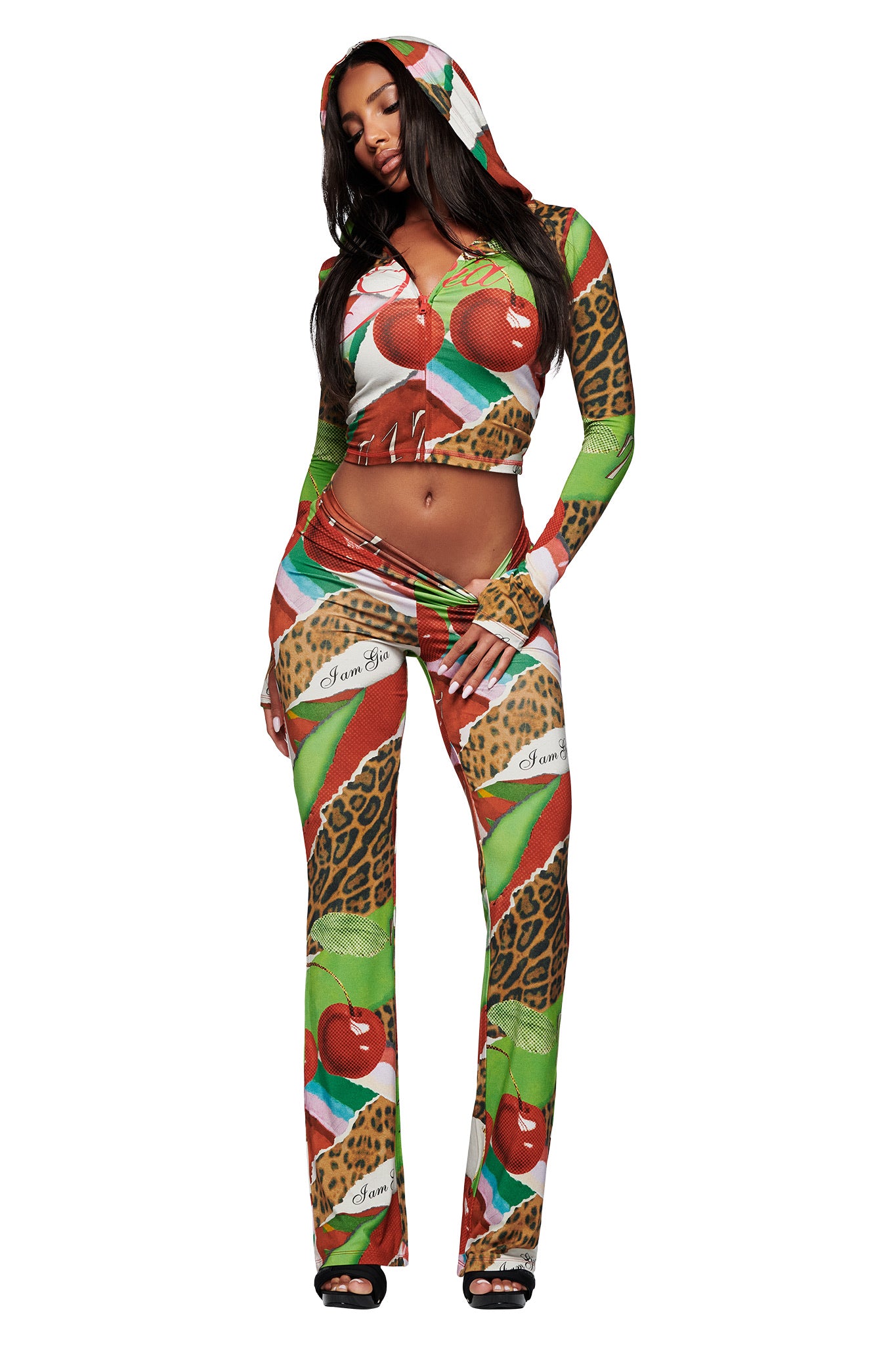A woman models the I.AM.GIA BLARE TRACKPANT - WILD CHERRY set, a vibrant two-piece featuring cherries, animal print, and cursive accents. The outfit includes a cropped zip-up hoodie and matching pants against a white background.
