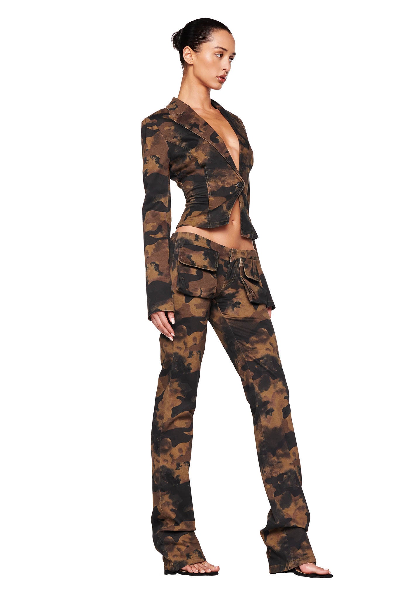 A model wearing the KIMANI PANT - GREEN : CAMO from I.AM.GIA, paired with a matching jacket. The cropped jacket is buttoned in the center, revealing a hint of midriff, while the low-rise pants feature large pocket details. The model's hair is styled in a sleek bun.