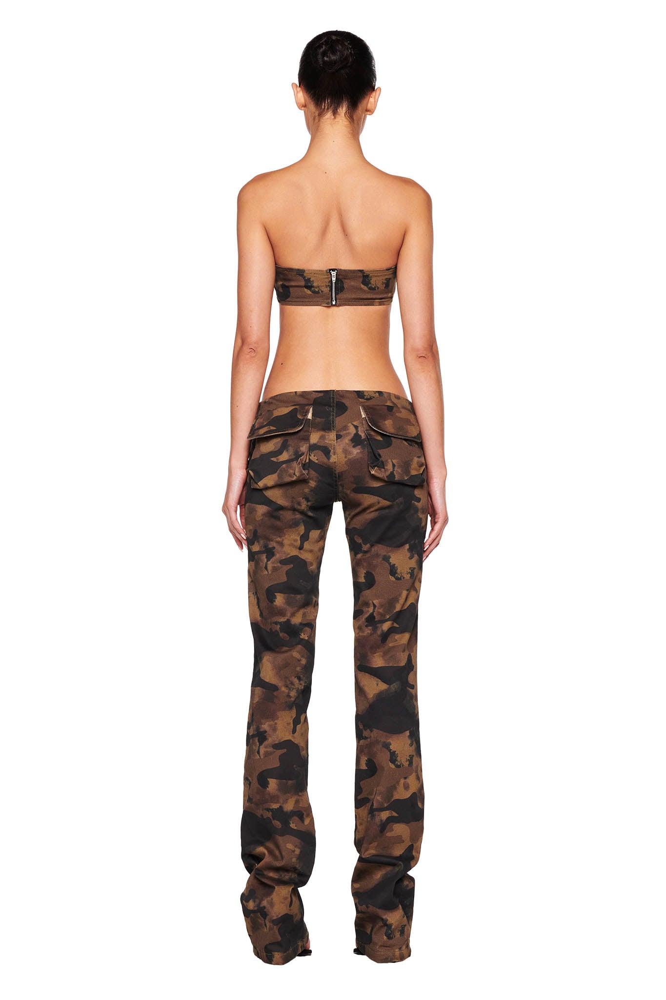 Rear view of a person standing and wearing an I.AM.GIA camo bandeau top and the KIMANI PANT - GREEN : CAMO. The top features a back zip closure, while the low-rise pants have a loose fit. The person's hair is tied up in a bun.