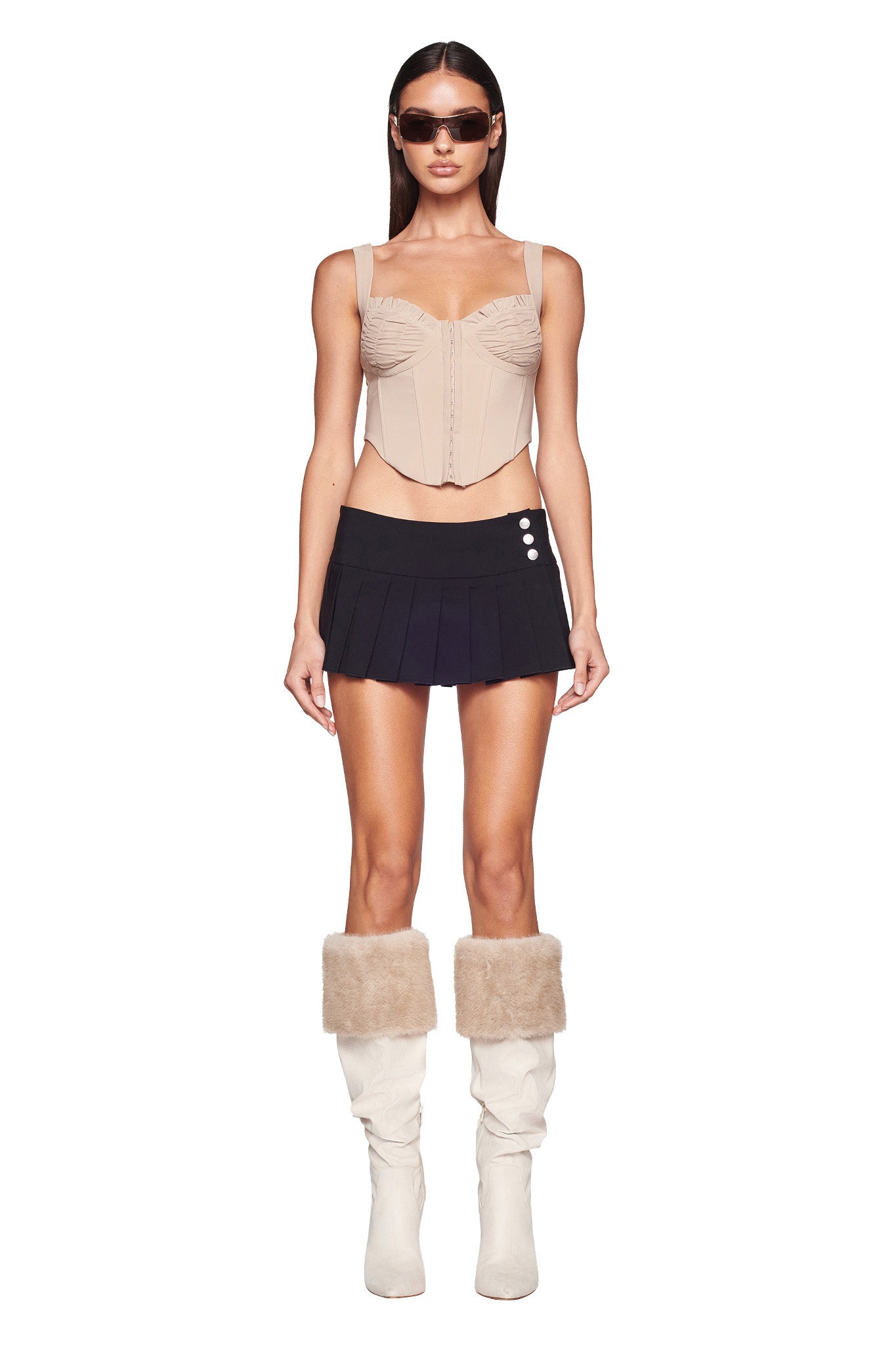 A woman stands modeling while wearing I.AM.GIA sunglasses, the BRI CORSET - NEUTRAL : TAN, and a navy pleated mini skirt with button detailing. She pairs the outfit with white knee-high boots featuring tan faux fur trim. The background is plain white, showcasing the ensemble.
