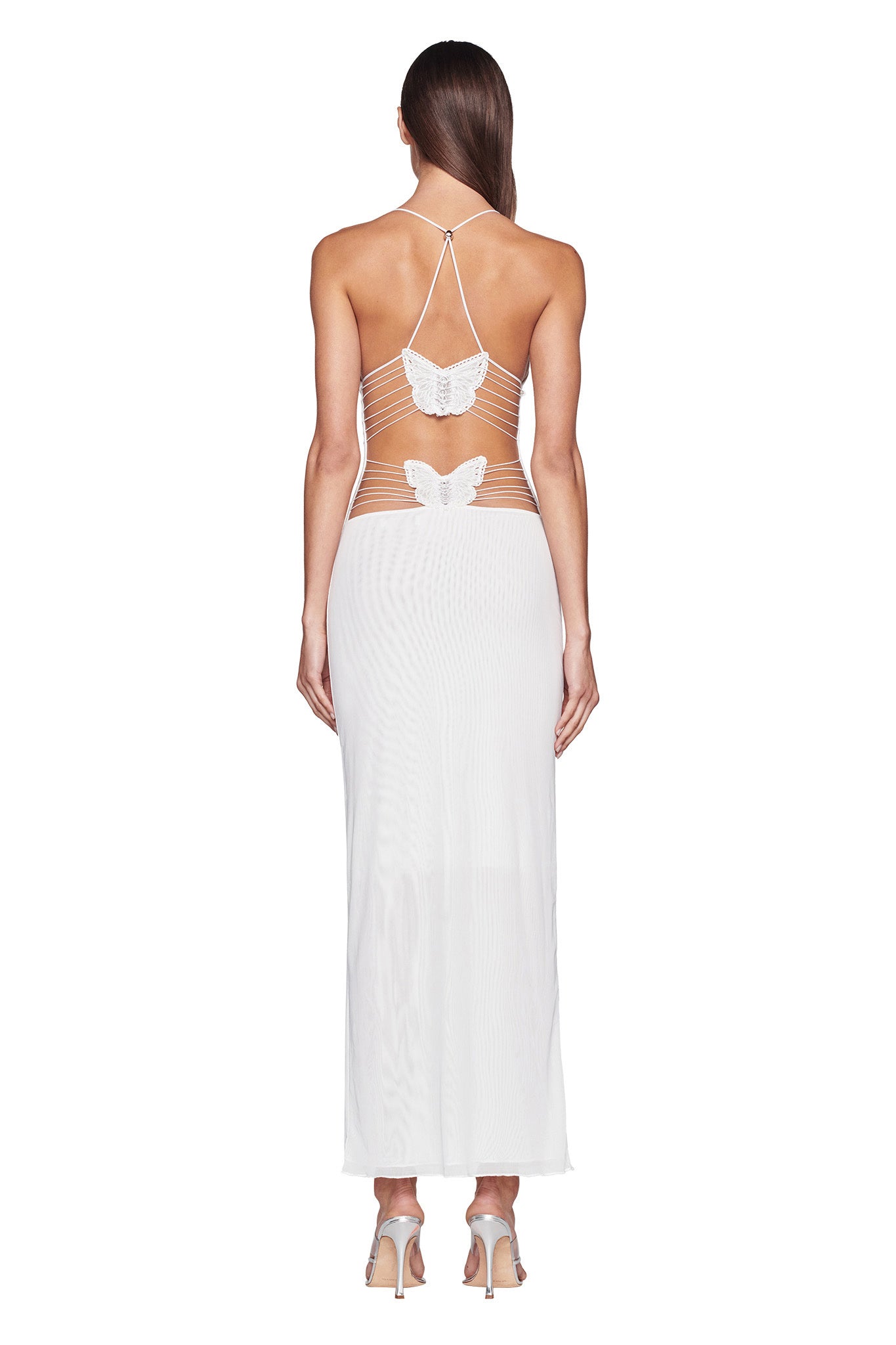 A woman is standing with her back to the camera, wearing the I.AM.GIA Ellery Dress in white. The dress is long and sheer with a strappy, crisscross back design featuring two butterfly-shaped embellishments. She has long, straight hair and is wearing high-heeled shoes.