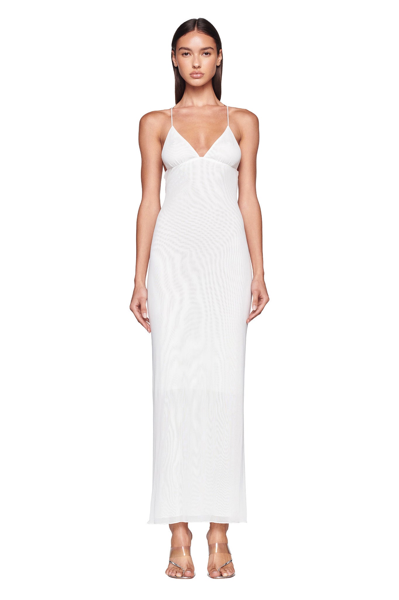 A woman with long, dark hair stands facing forward in a full-length, sleeveless ELLERY DRESS - WHITE by I.AM.GIA, featuring thin straps. She appears calm and composed, complemented by transparent high-heeled sandals. The background is white and plain.