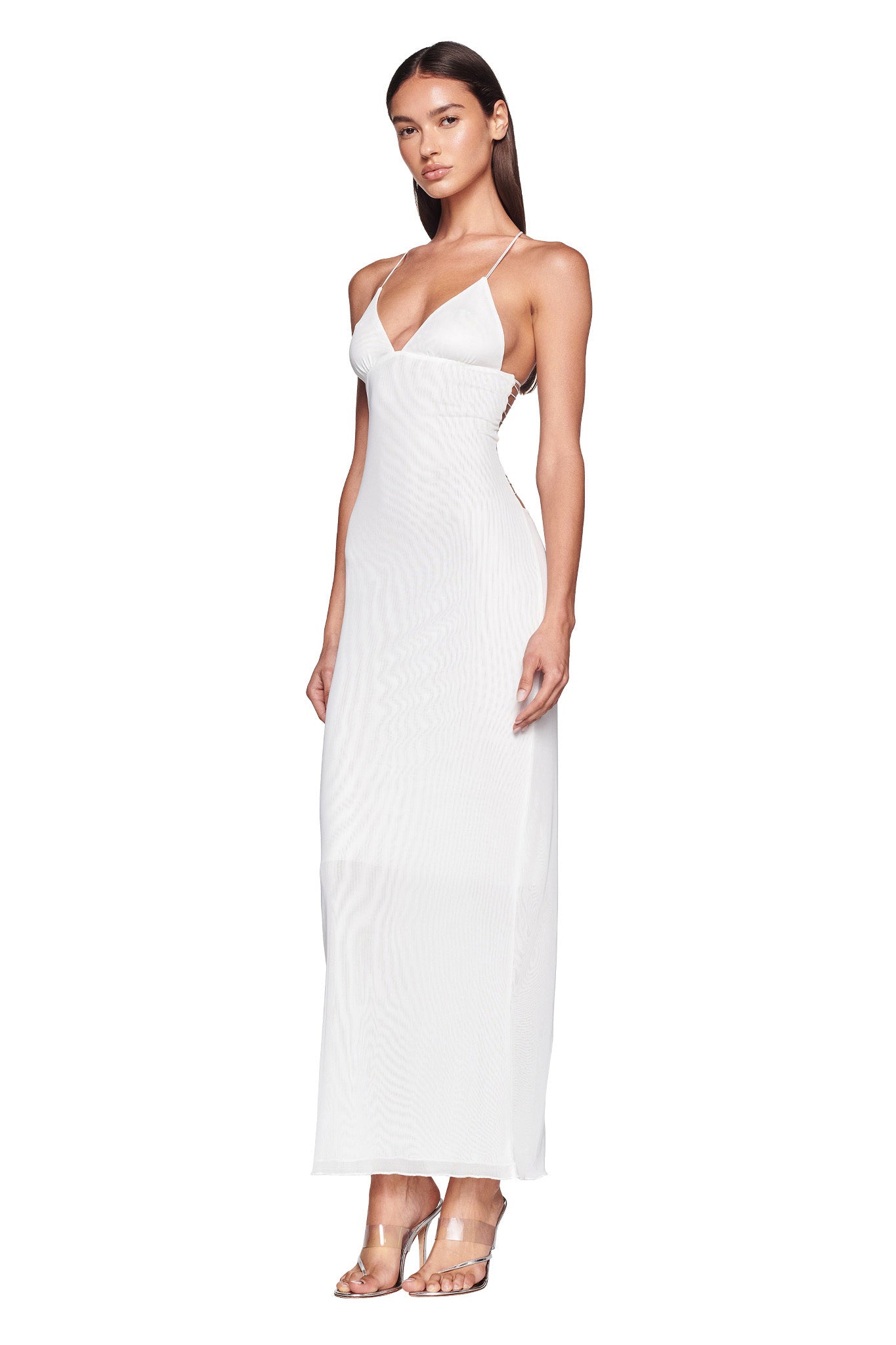 A woman in a white, form-fitting, sleeveless ELLERY DRESS by I.AM.GIA stands in front of a plain white background. She is wearing high-heeled, transparent sandals. Her pose is relaxed with one hand resting by her side and the other slightly behind her back.