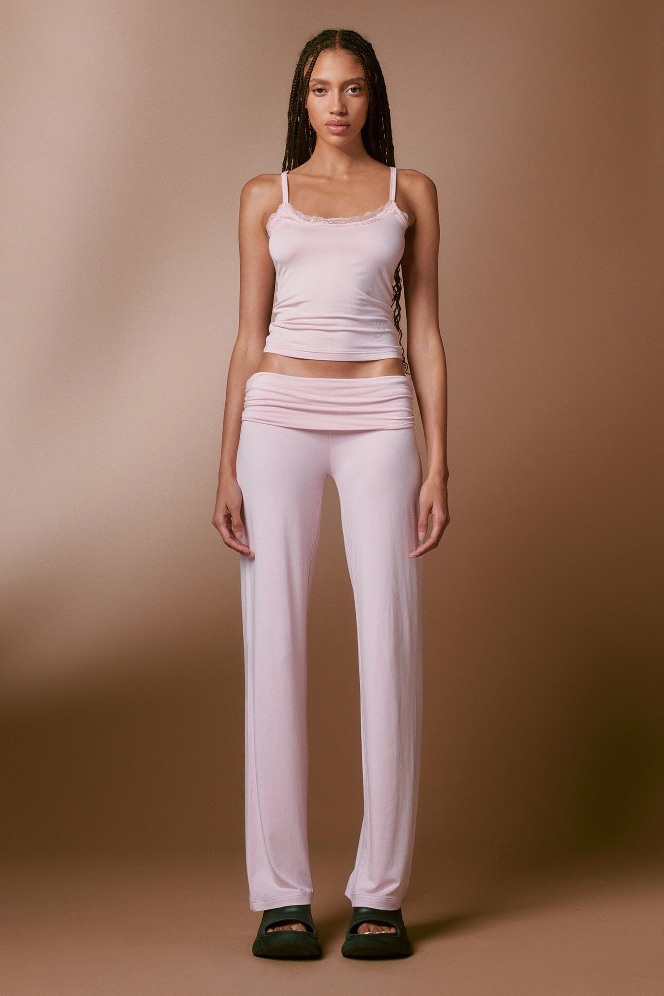 A person with long braids is standing against a plain background, wearing the cozy I.AM.GIA ERIN TOP in pink along with matching loose bamboo-fabrication pants. They are also sporting black closed-toe footwear. The overall color scheme is soft and neutral.