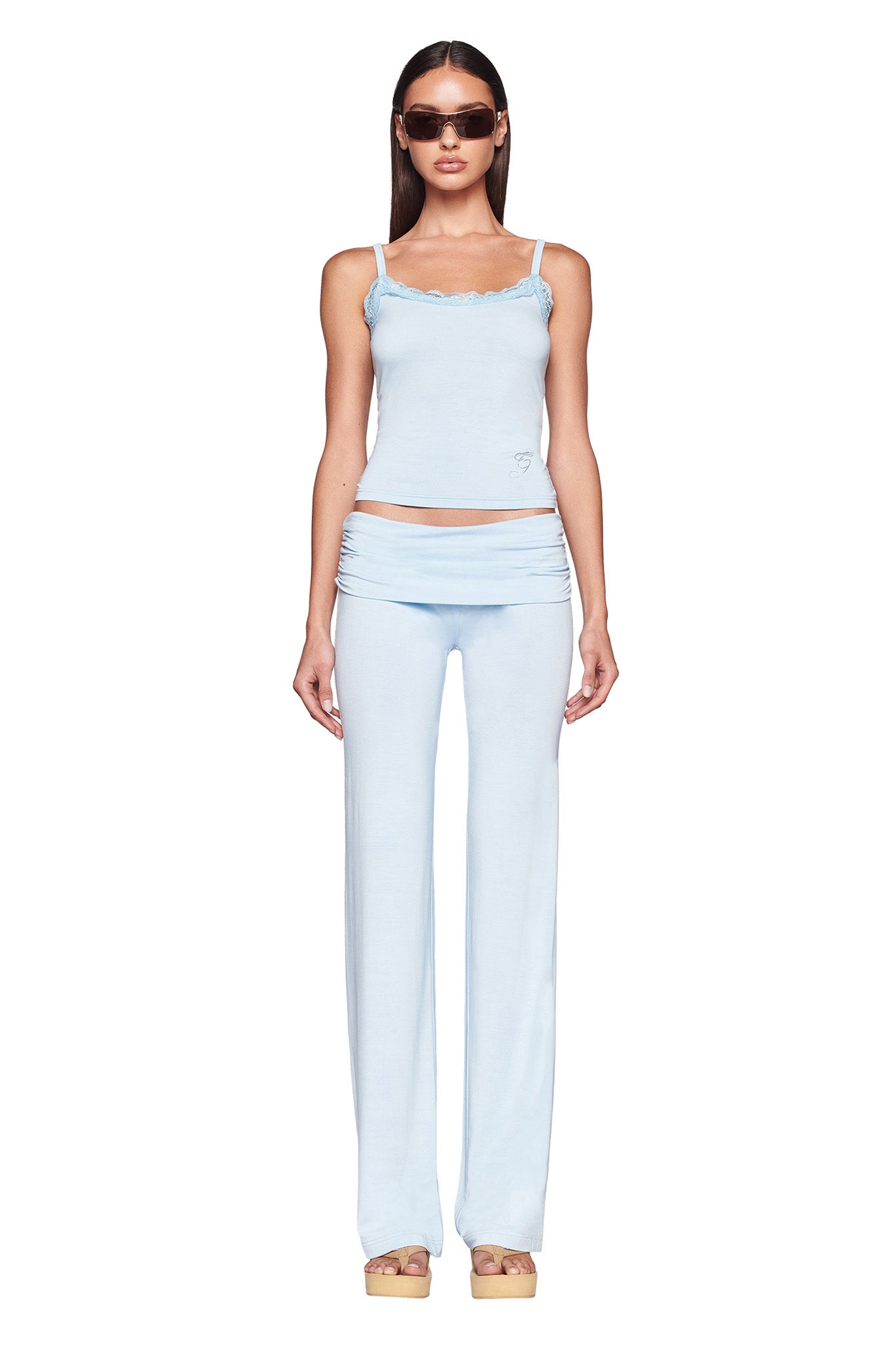 A woman stands facing forward, wearing a light blue, sleeveless "ERIN TOP - BLUE : BABY BLUE" by I.AM.GIA with ruffled edges and matching low-rise pants. She has long, dark hair and is wearing brown sunglasses. She also wears tan platform sandals. The background is completely white.