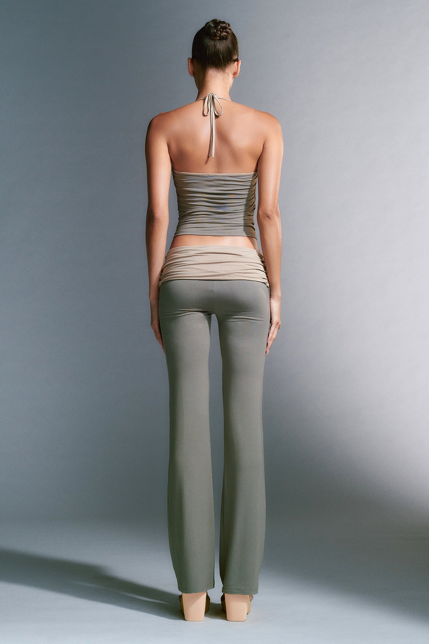 A person with a bun hairstyle is seen from the back, wearing an I.AM.GIA form-fitting, backless halter top and high-waisted, wide-leg JANNIS PANT in green. The backdrop is plain and well-lit.