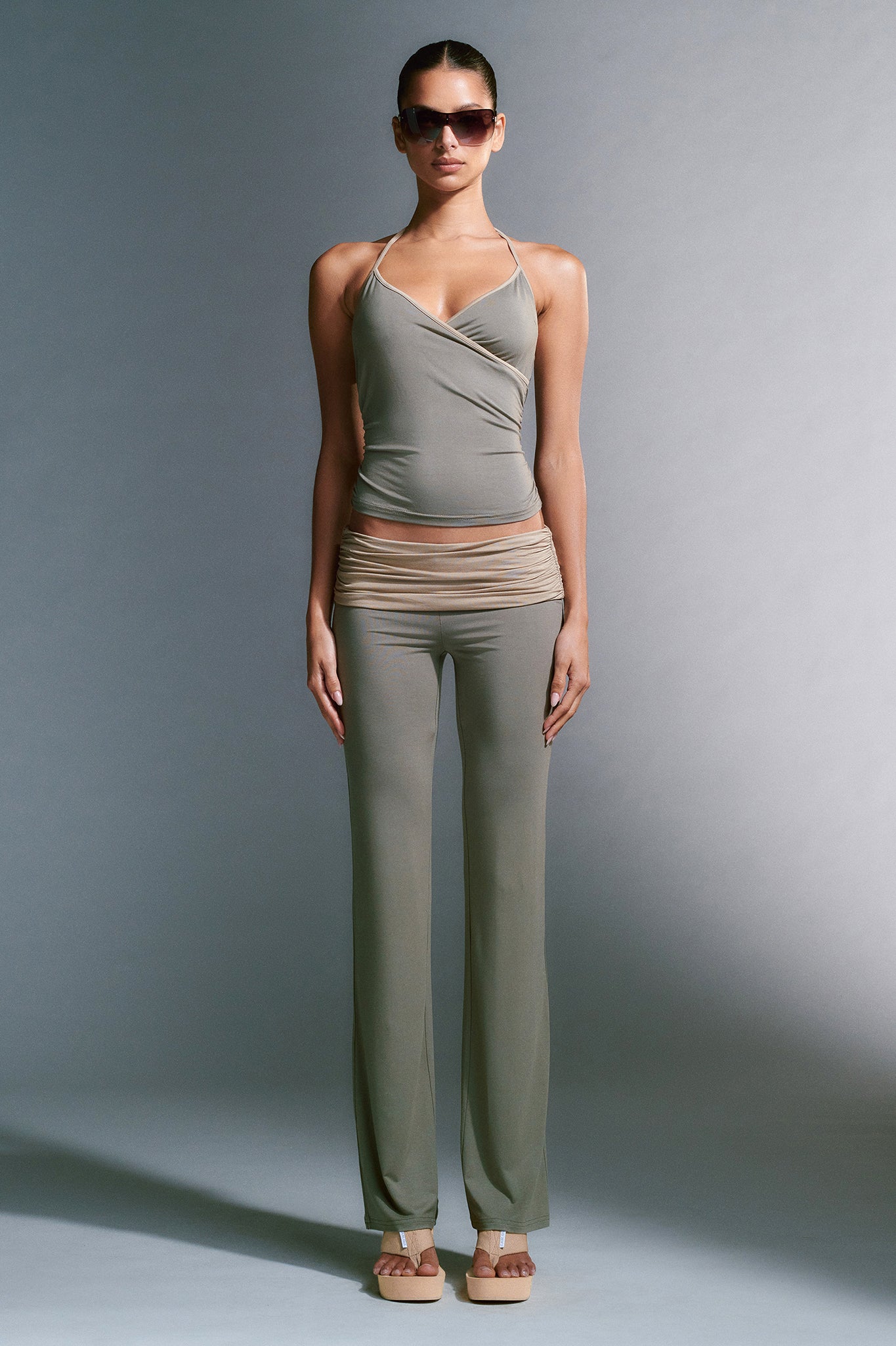 A person stands confidently against a neutral background, wearing a fitted, asymmetrical Jannis Tank in green by I.AM.GIA paired with matching pants. They accessorize with large, dark sunglasses and beige platform sandals. Their hair is pulled back, and their arms rest at their sides.