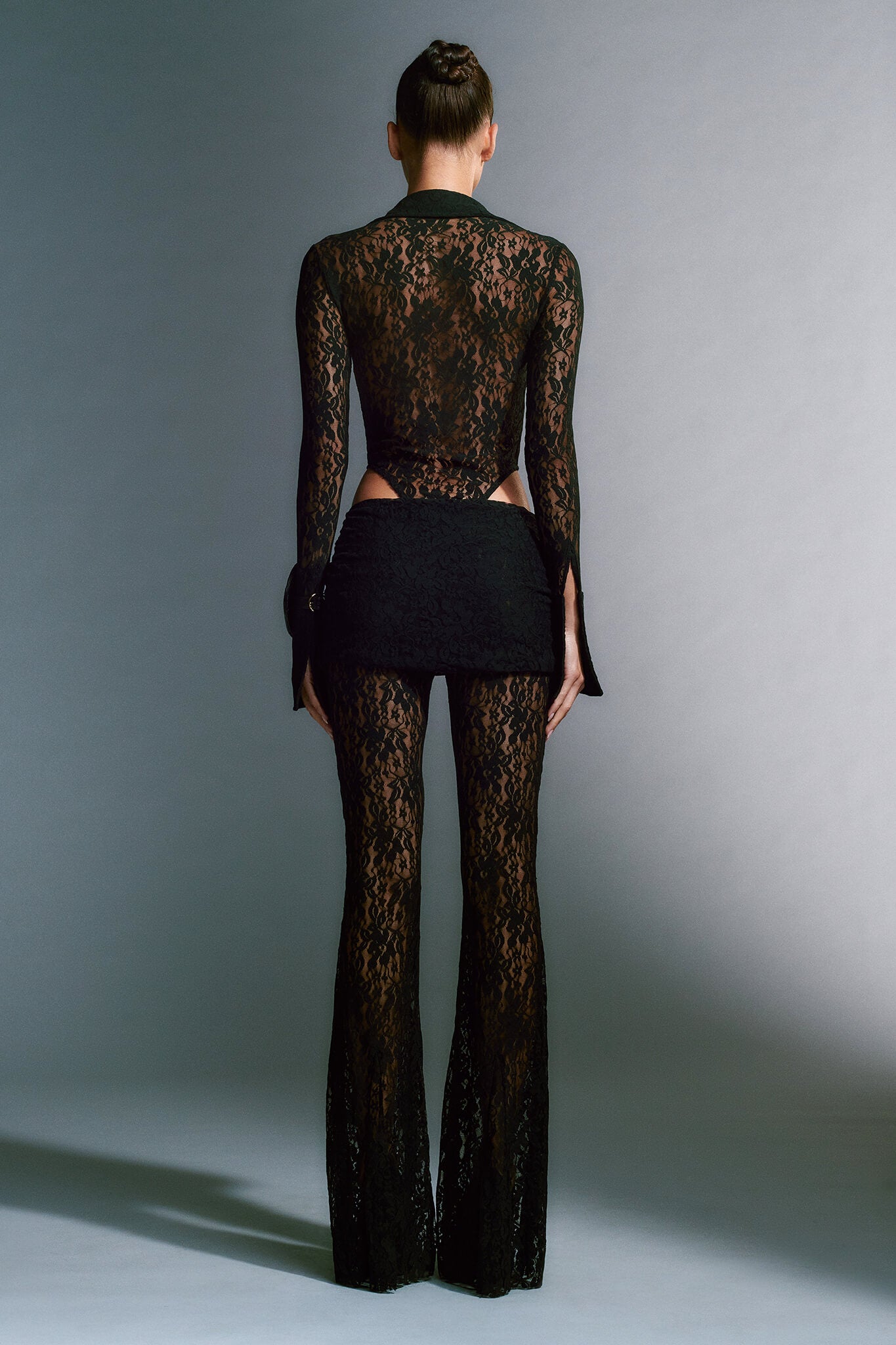 A model stands facing away, showcasing the KYLEAH PANT - BLACK : LACE from I.AM.GIA. The form-fitting ensemble includes long sleeves, black lace pants with a flared leg silhouette, and a short skirt-like layer around the low-rise waistline. The background is a neutral gray gradient.