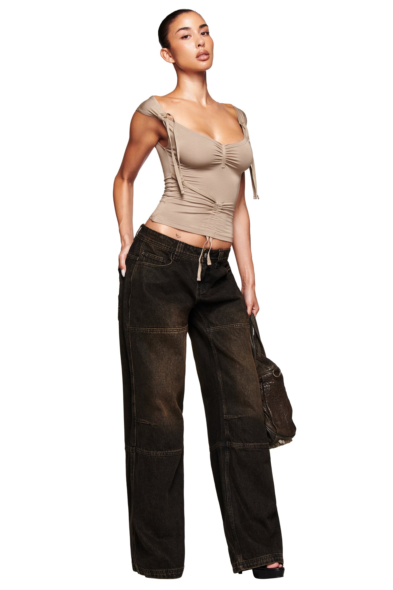 A woman stands confidently against a white background, wearing I.AM.GIA MISTY JEAN - BLACK paired with a beige ruched cap sleeve top and holding a bag at her side.