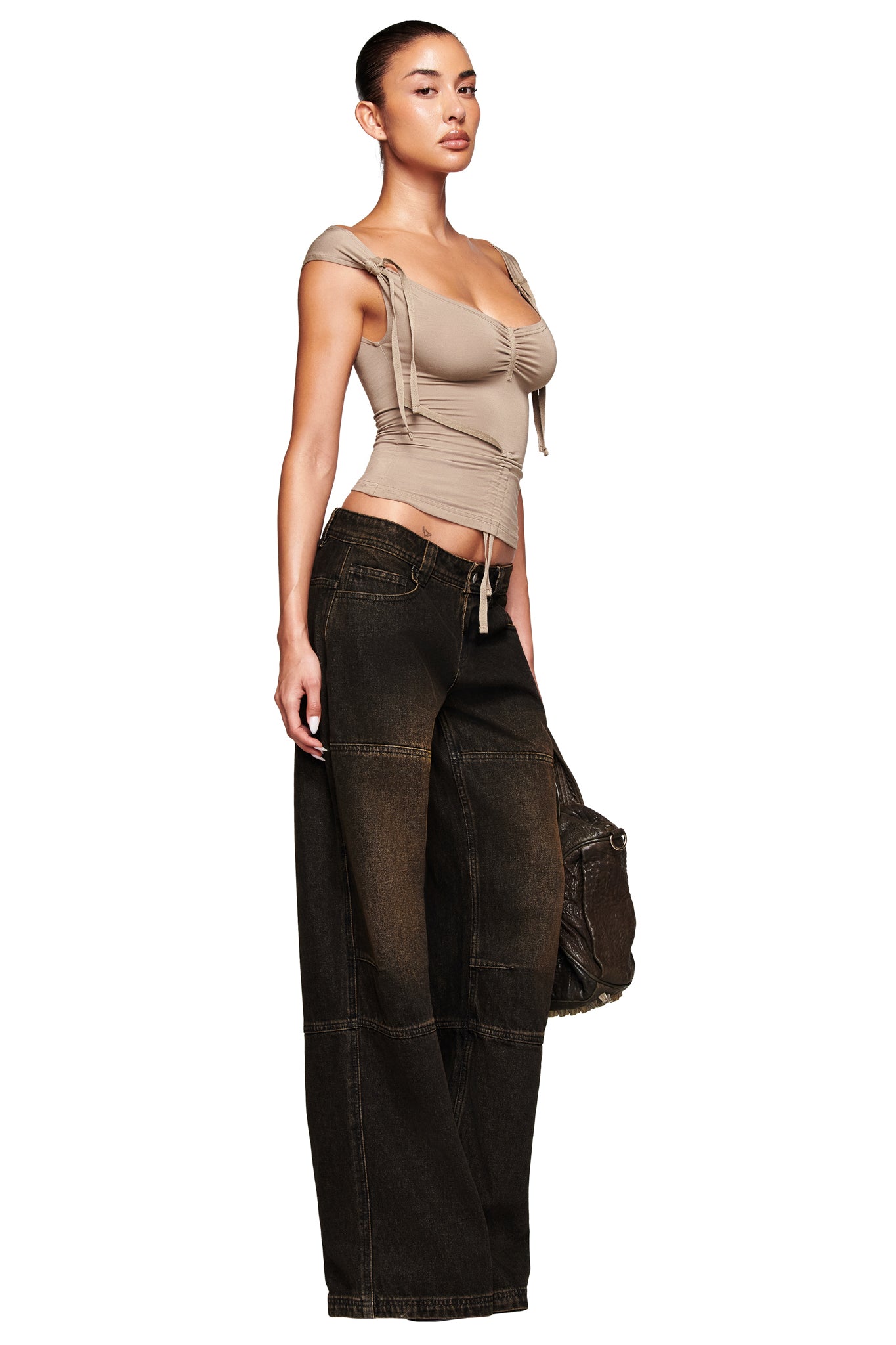 A person stands confidently in a fitted beige ruched top and loose-fitting, low-rise MISTY JEAN - BLACK by I.AM.GIA. They carry a dark bag and have their hair styled in a sleek bun.