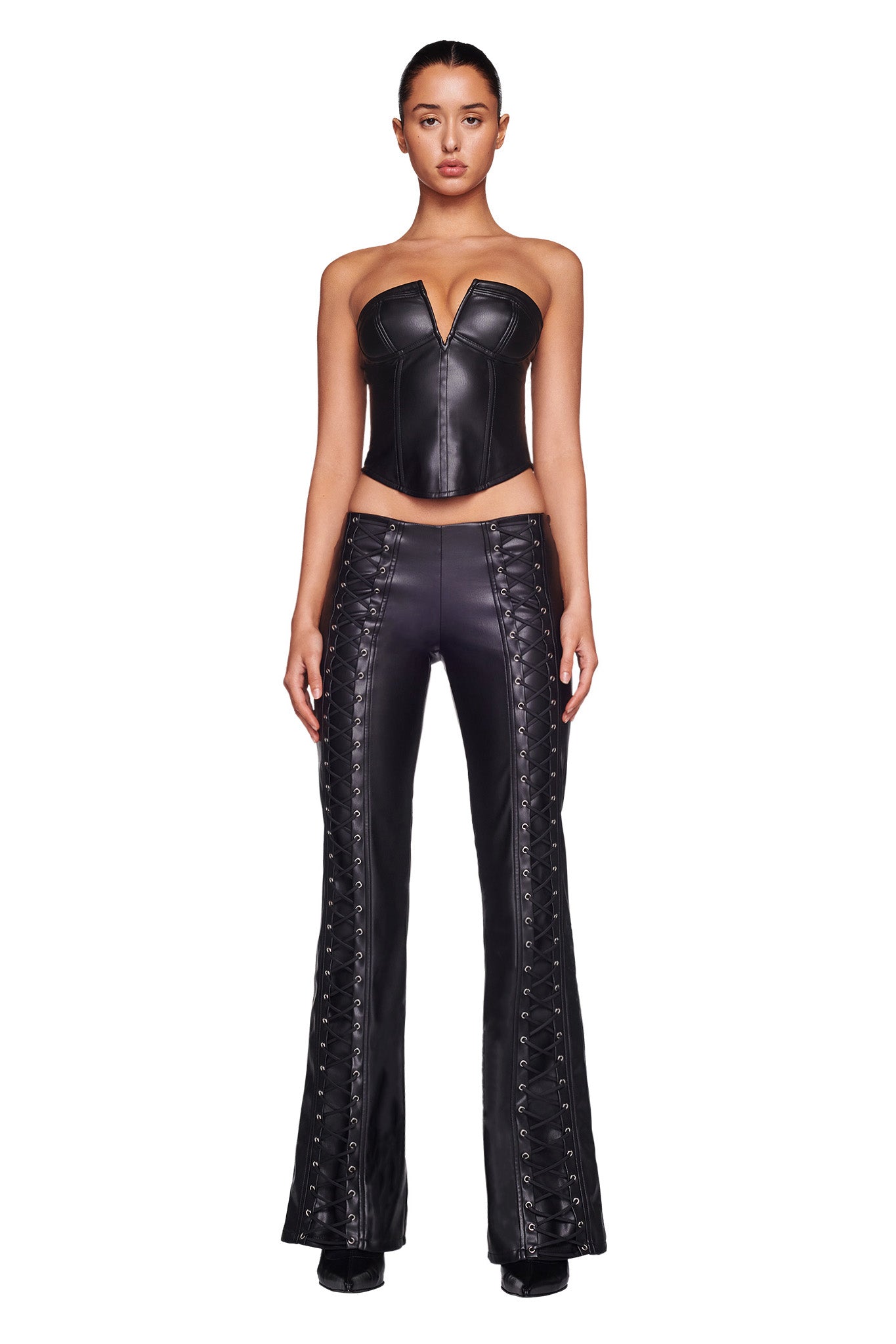 A woman stands facing forward wearing the EVANNA CORSET - BLACK, a black, strapless leather top with a V-shaped neckline from I.AM.GIA, paired with matching black leather pants featuring lace-up detailing along the sides. Her hair is pulled back, and she is standing against a plain white background.