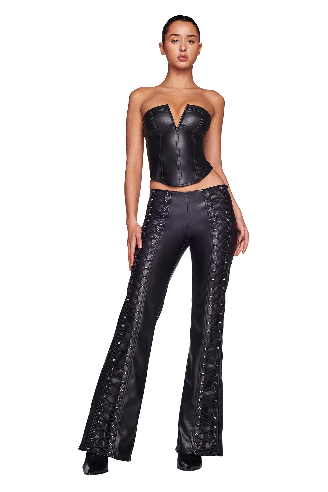 A person stands confidently in the EVANNA CORSET - BLACK from I.AM.GIA, a strapless leather top with a deep neckline. They are also wearing matching black leather pants that feature lace-up detailing along the sides. Against a plain white background, their pose exudes style and poise.