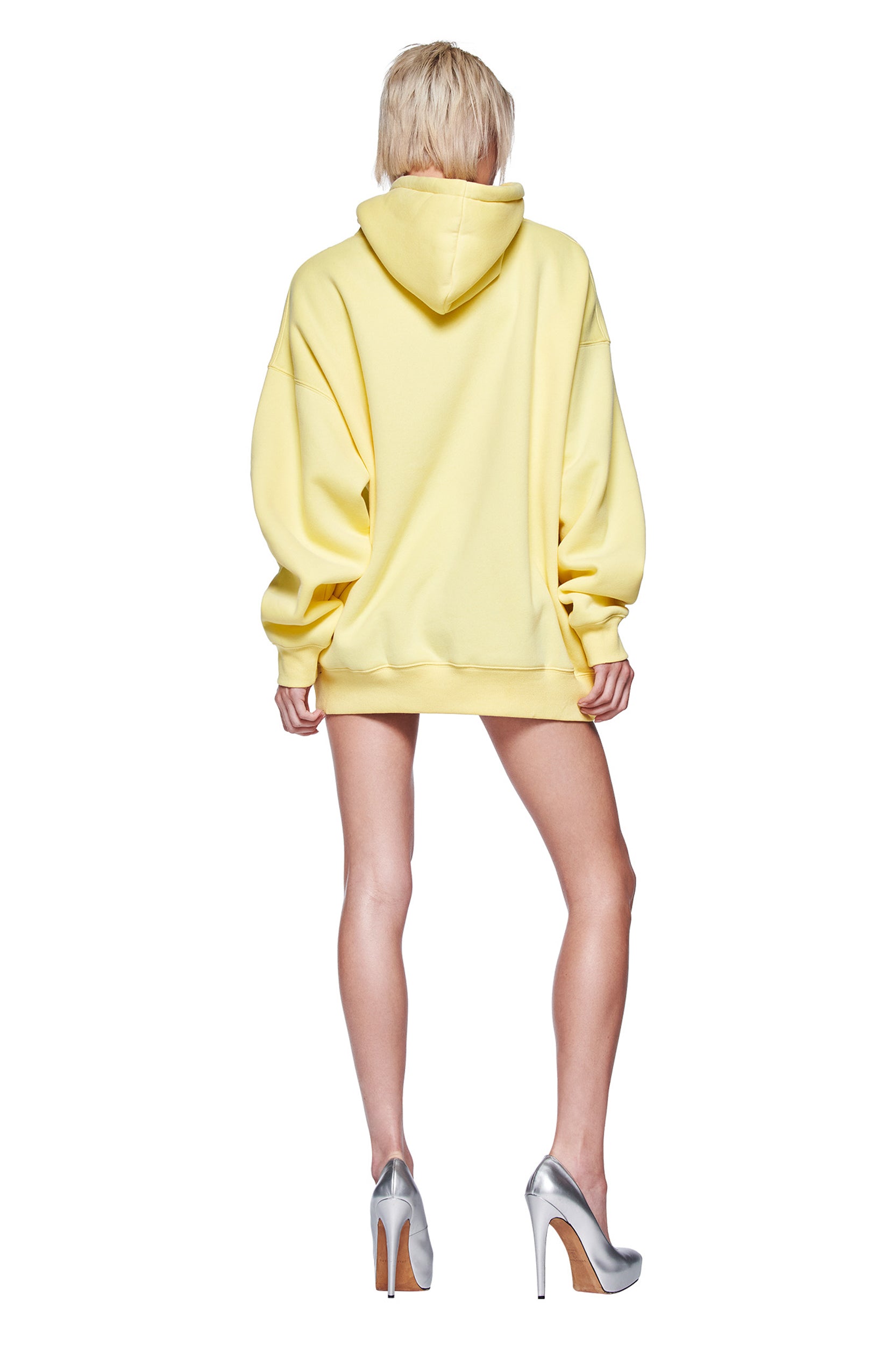 A person with short blonde hair faces away from the camera, wearing the I.AM.GIA DORIAN HOODIE in yellow and silver high heels. The plain white background makes the I.AM.GIA branding on the hoodie stand out.