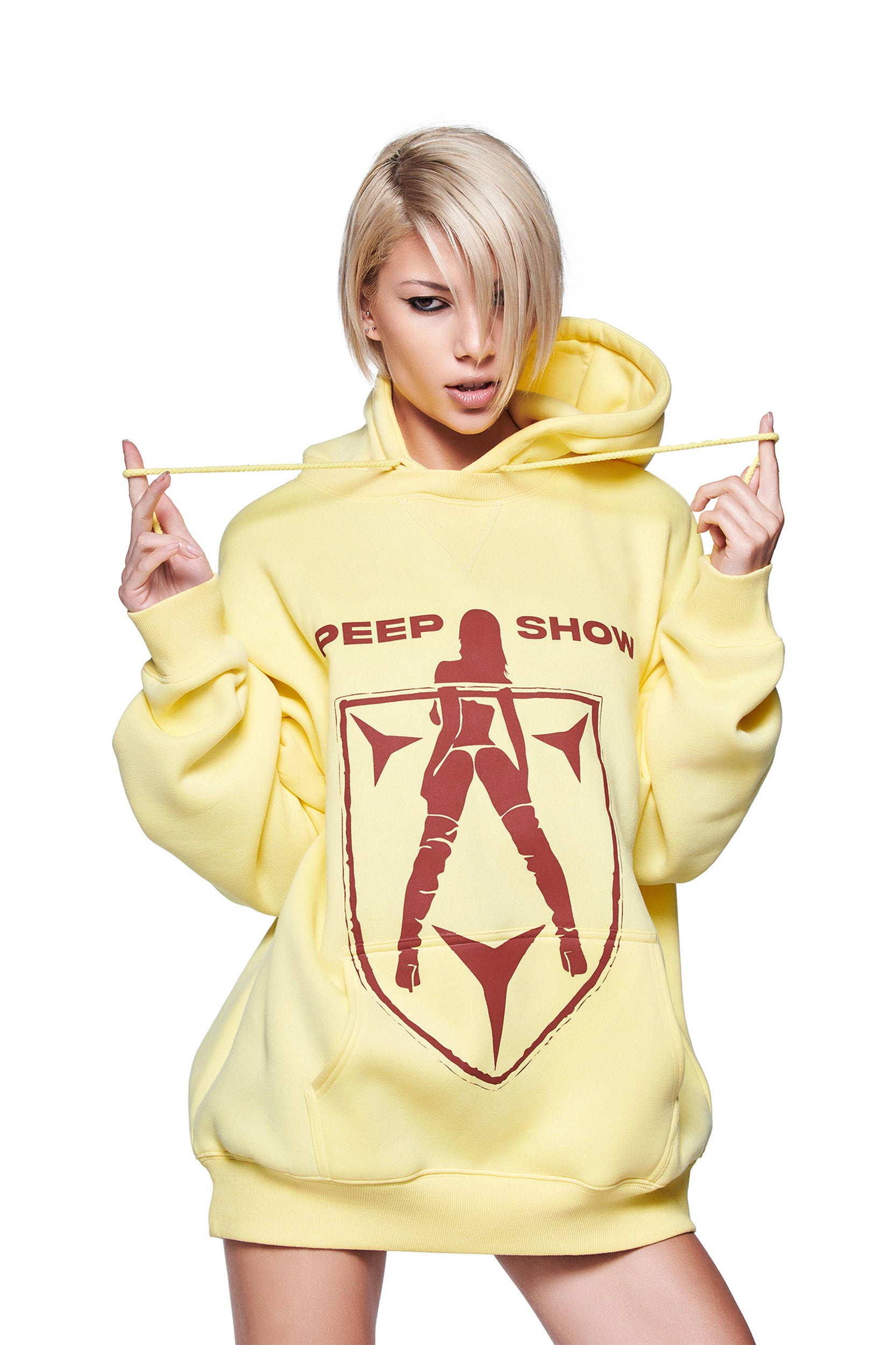 A person with short blonde hair wears the I.AM.GIA DORIAN HOODIE in yellow, featuring an oversize silhouette, "PEEP SHOW" text, and a woman-in-boots graphic. They hold the hoodie strings while standing against a plain white background.