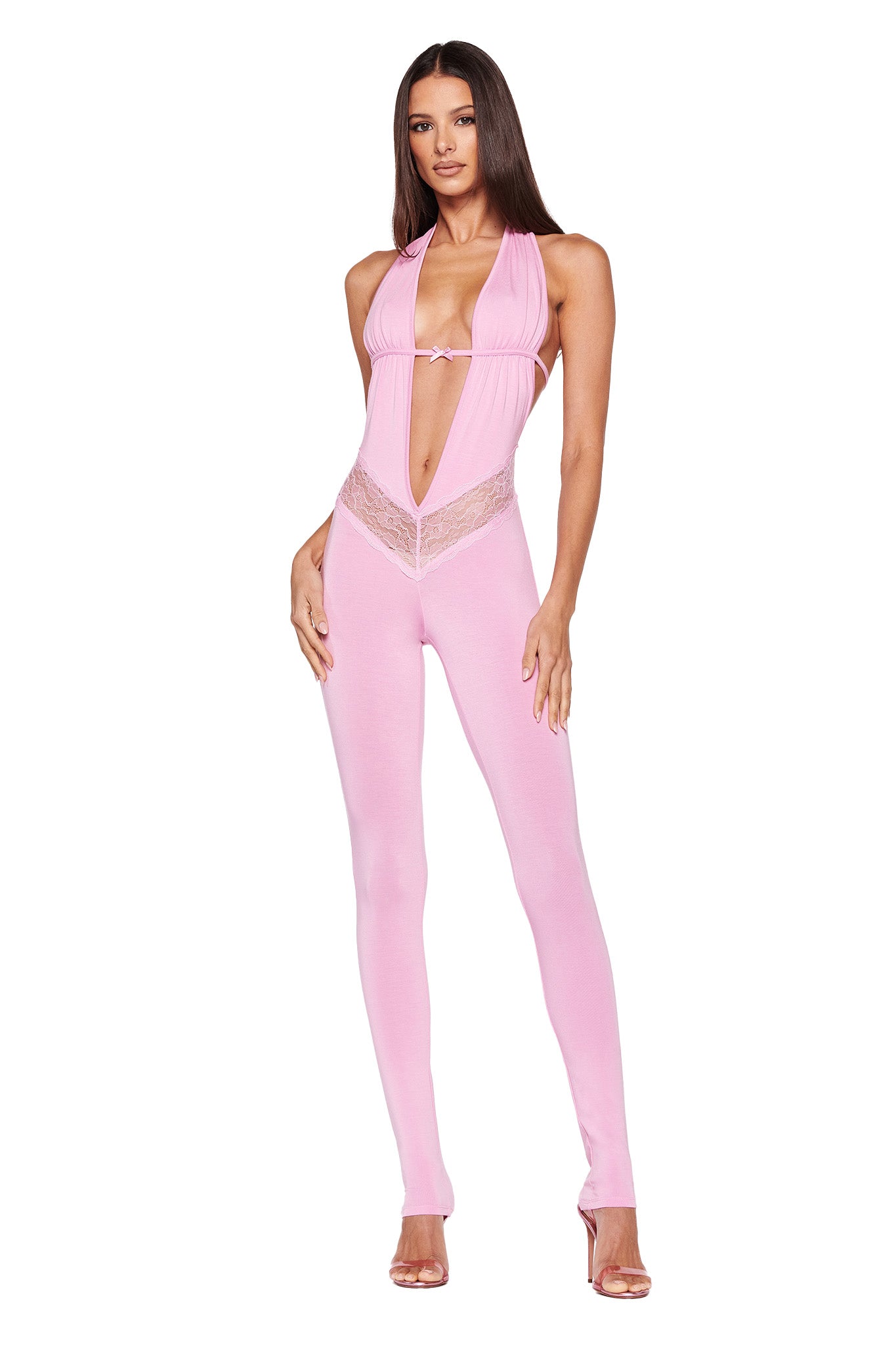 A woman with long straight hair is wearing the I.AM.GIA ASTORA JUMPSUIT in pink, featuring a halter-neck design, a fitted silhouette, a deep V neckline, and lace detailing at the waist. She stands with one hand on her hip and the other resting by her side, posing against a plain white background.