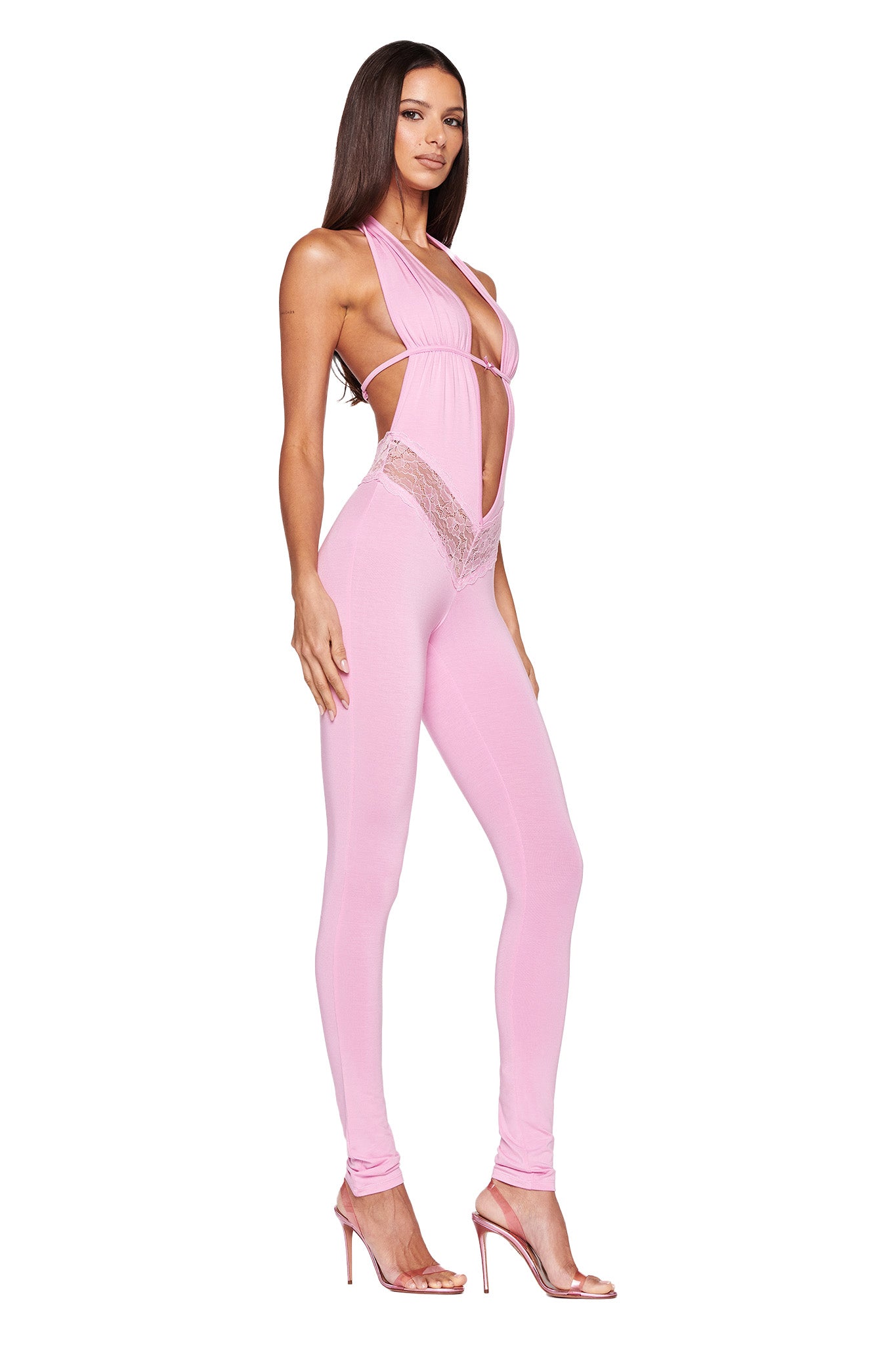 A woman is standing in the ASTORA JUMPSUIT - PINK by I.AM.GIA, which features cut-out details on the sides and lace accents around the waist. The outfit includes a halter-style top with a plunging neckline. She is wearing high-heeled sandals and has long, straight hair.