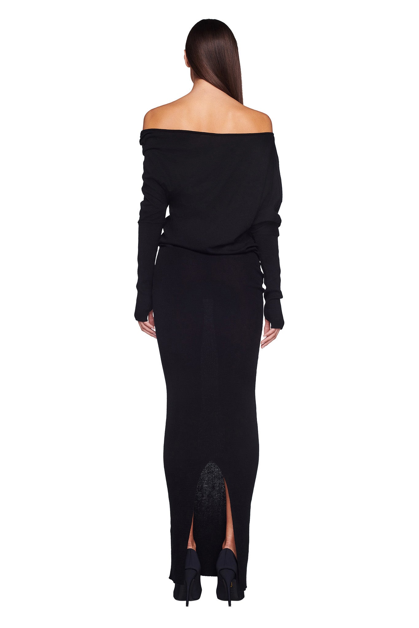 A woman with long brown hair stands facing away, donning the I.AM.GIA GISELLE MAXI DRESS in black, which extends to her ankles and features a slit at the back hem. She complements her outfit with black high-heeled shoes.