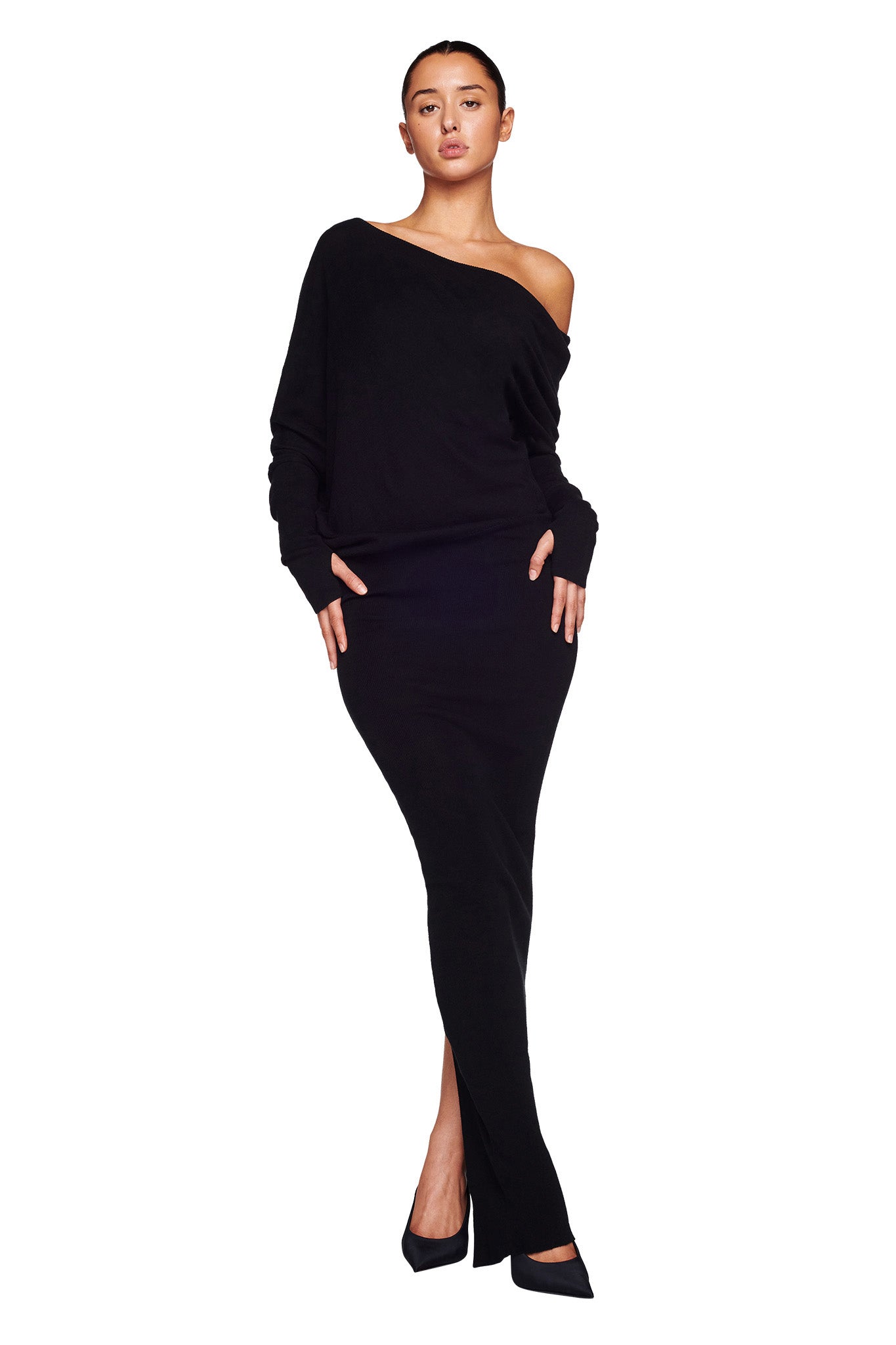 A person stands against a plain white background, wearing the sophisticated GISELLE MAXI DRESS - BLACK from I.AM.GIA, featuring an off-the-shoulder neckline. One hand rests in a pocket while they wear black pointed-toe shoes. The long, fitted dress accentuates a sleek silhouette.