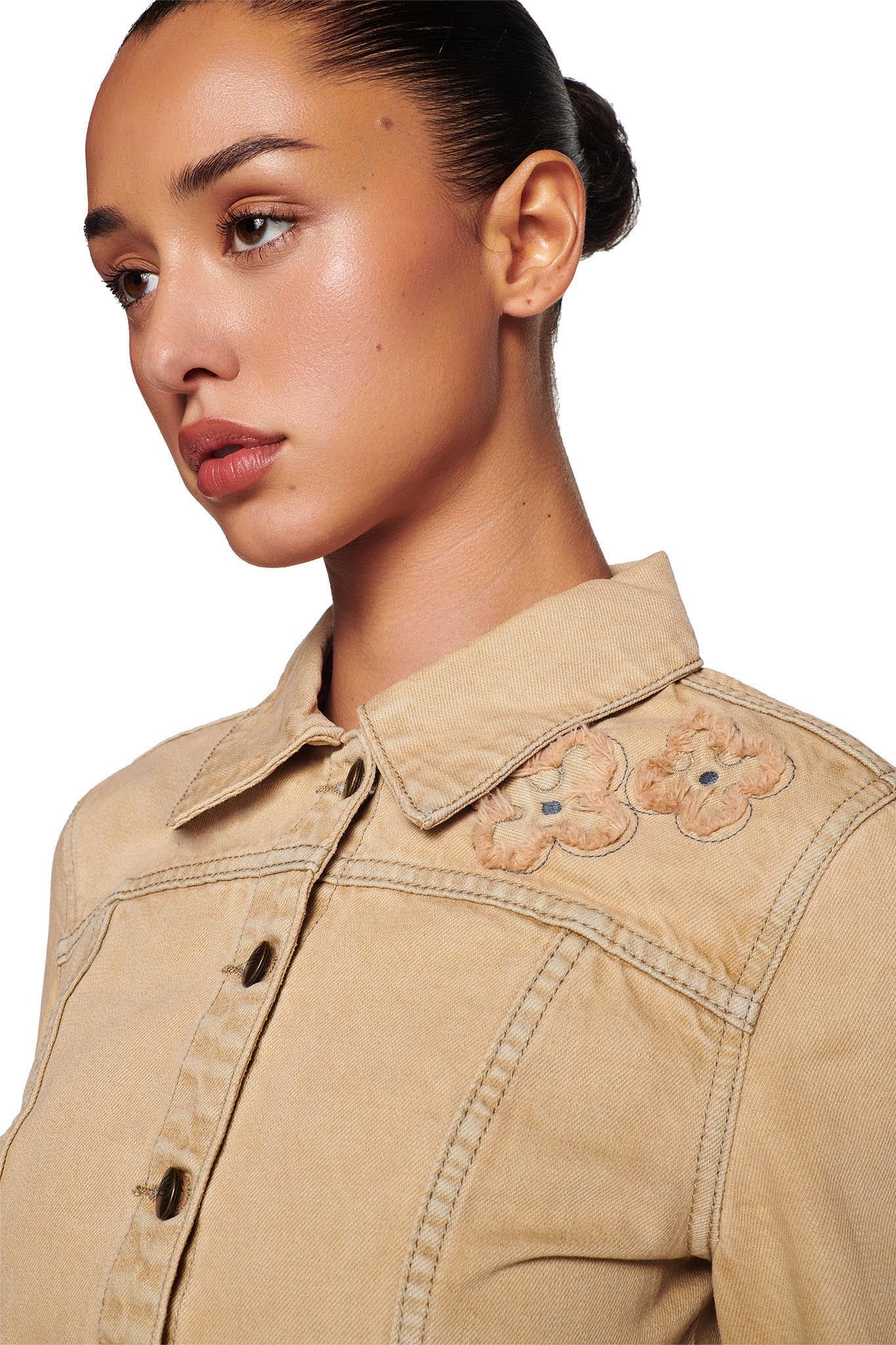 A woman with a bun is wearing an I.AM.GIA FAUNA JACKET - BEIGE : TAN DENIM, which features delicate floral embroidery near the shoulders. The image is cropped to show her from the chest up, and she gazes to the side against a plain white background.
