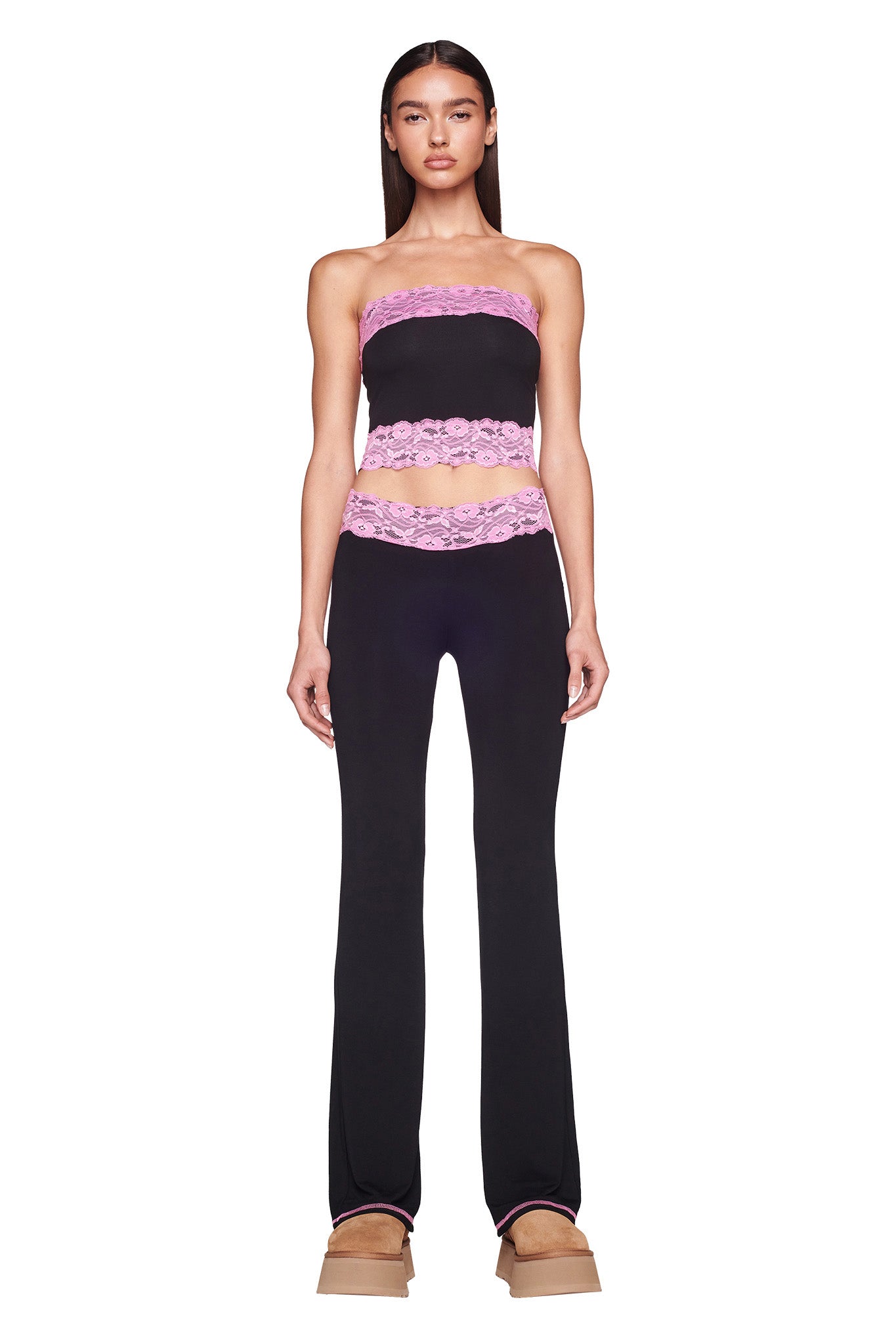 A woman stands facing forward, wearing the chic I.AM.GIA TARSHA BOOB TUBE in black with pink lace details at the top and bottom, paired with matching black pants featuring pink lace at the waistband. She has straight hair and is wearing light brown platform shoes.