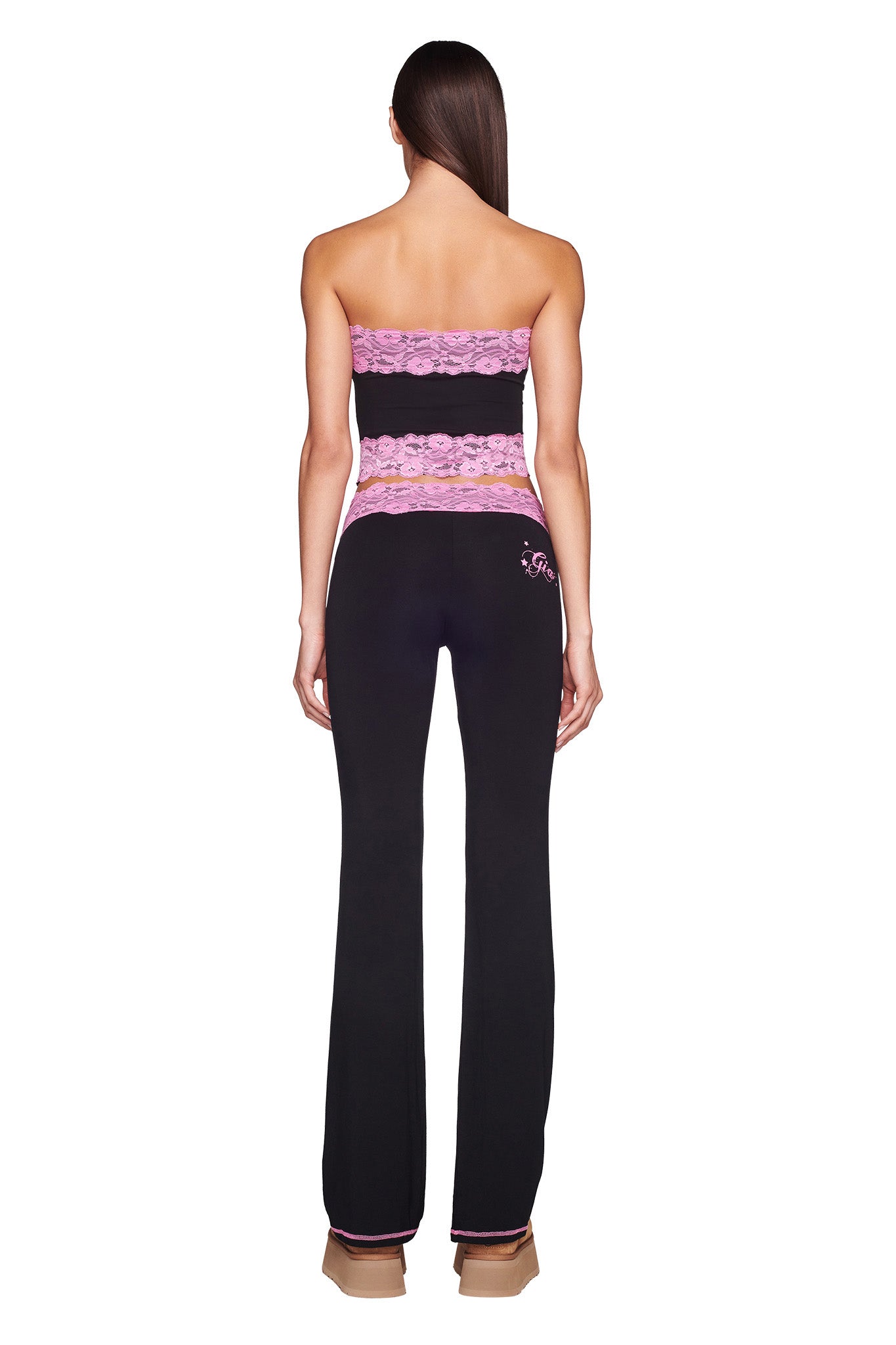 A woman is standing with her back facing the camera, wearing the TARSHA BOOB TUBE in black and pink from I.AM.GIA, coupled with matching black pants. She has long straight dark hair, and her outfit emphasizes her slender figure against a plain white background.