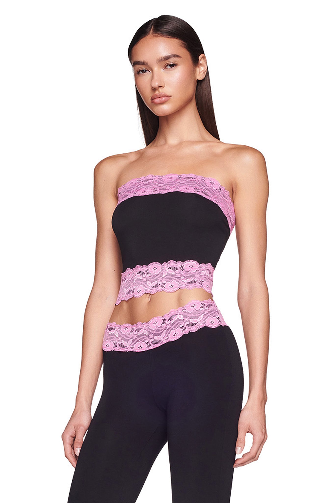 A woman with long dark hair stands against a white background, wearing the TARSHA BOOB TUBE - BLACK/PINK and matching black leggings from I.AM.GIA. The top and leggings feature pink lace trim around the edges. She is looking directly at the camera with a neutral expression.