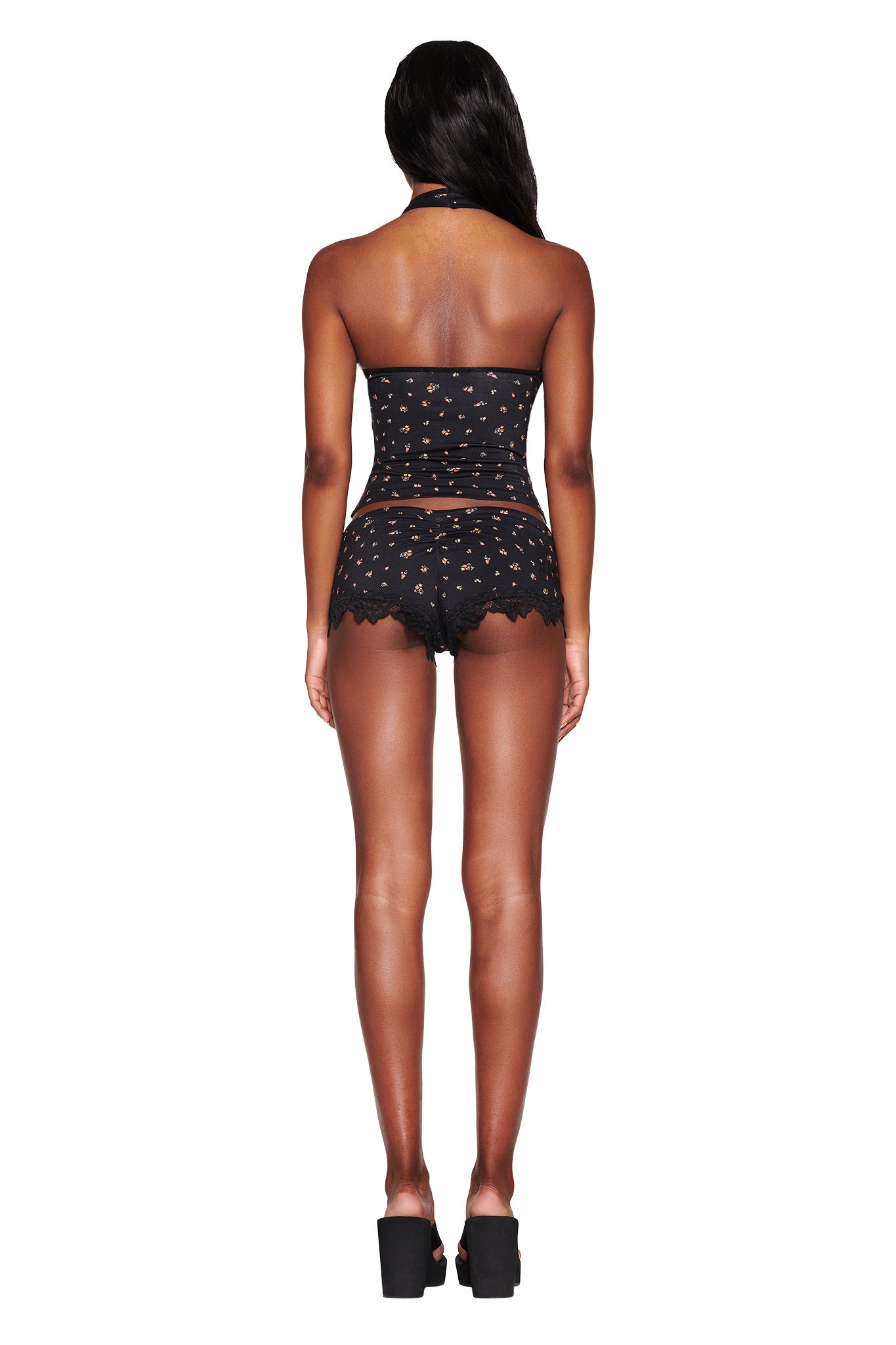 A person models I.AM.GIA's CELESTE SHORT - BLACK : DITSY FLORAL, featuring a strapless top and matching lace-edged shorts, standing on high platform shoes against a white background. The outfit elegantly accentuates a bodycon silhouette and their curves.