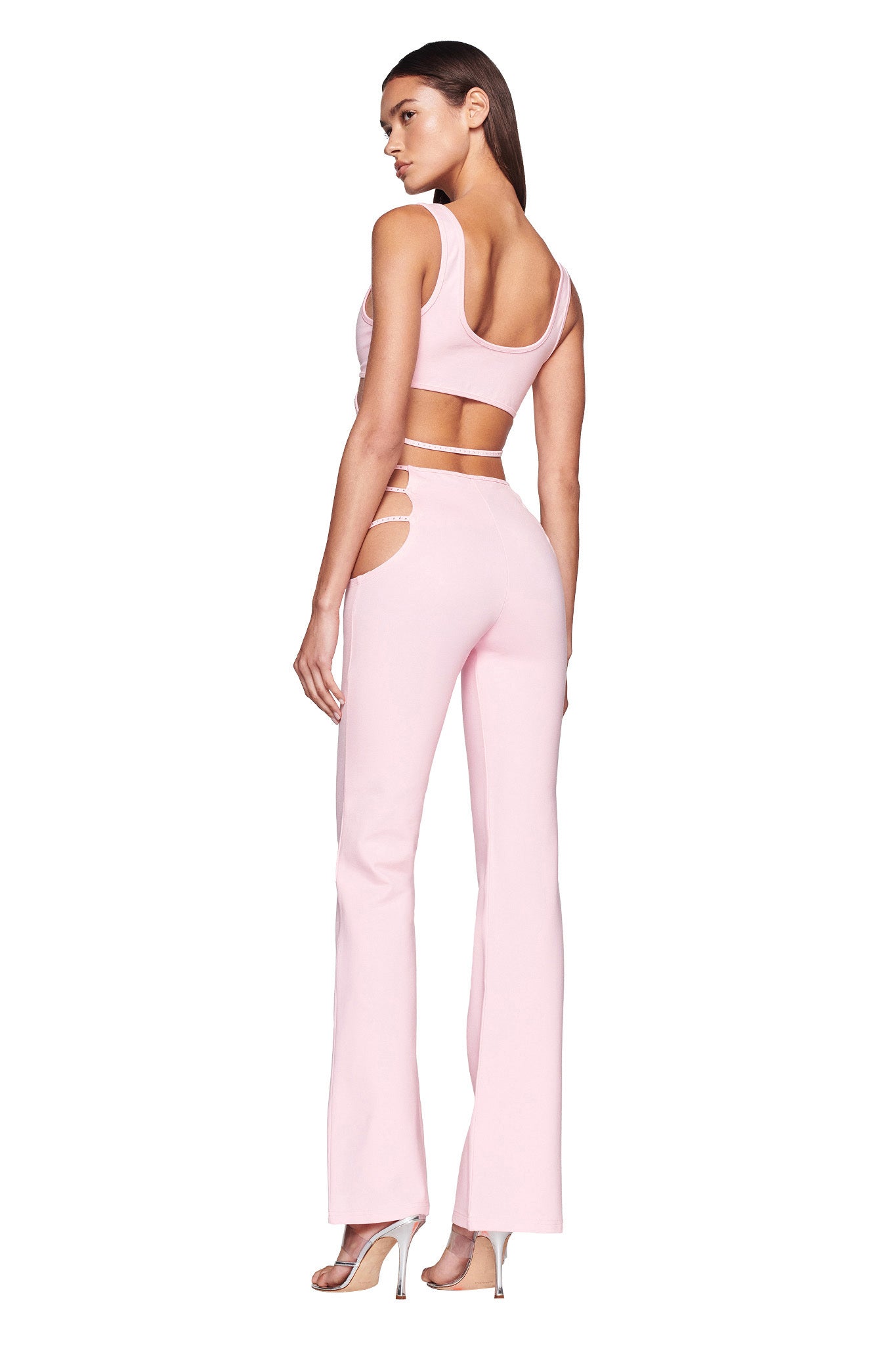 A person stands facing backward, showcasing an I.AM.GIA sleeveless, light pink crop top and the matching ROXANNA PANT in Baby Pink, featuring cutout details on the waist. They wear high-heeled shoes and have long, straight hair. The background is plain white.