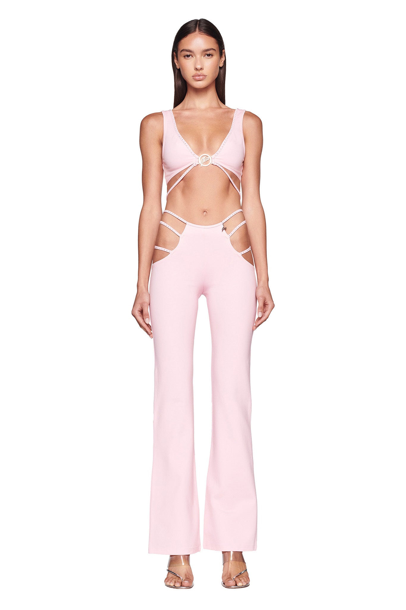 A woman stands against a plain white background wearing the I.AM.GIA ROXANNA PANT in Baby Pink, a jumpsuit featuring multiple cut-outs around the midriff and sides, flared legs, and a circular ring detail at the bust. She completes her look with clear high-heeled sandals.