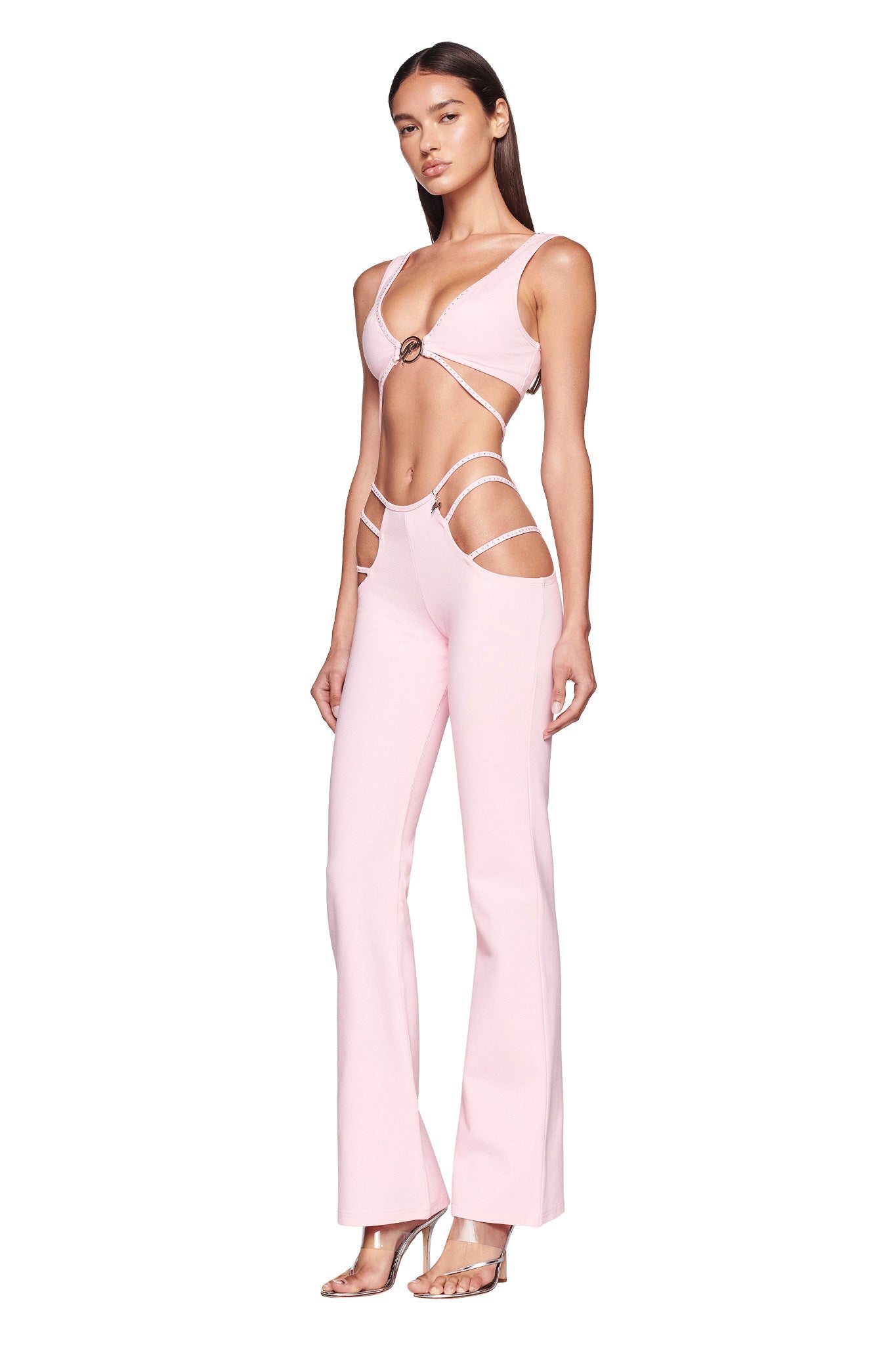 A woman stands wearing the ROXANNA PANT - PINK : BABY PINK from I.AM.GIA, a stylish two-piece outfit with cutouts on the sides of the top and pants, revealing her midriff and hips. The top features a mid-ring detail, and the pants have side cutout straps. She completes the look with clear high-heeled sandals.