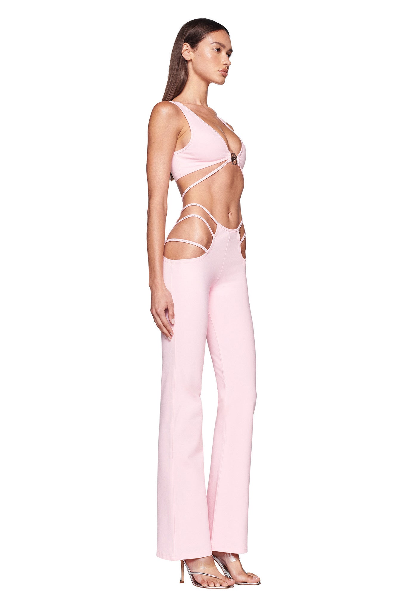 A woman is standing sideways in a light pink ROXANNA PANT - PINK by I.AM.GIA. The jumpsuit, which features cut-outs revealing her midriff and sides, has thin straps and bell-bottom legs. She is wearing high-heeled sandals and her long, straight hair cascades down her back.