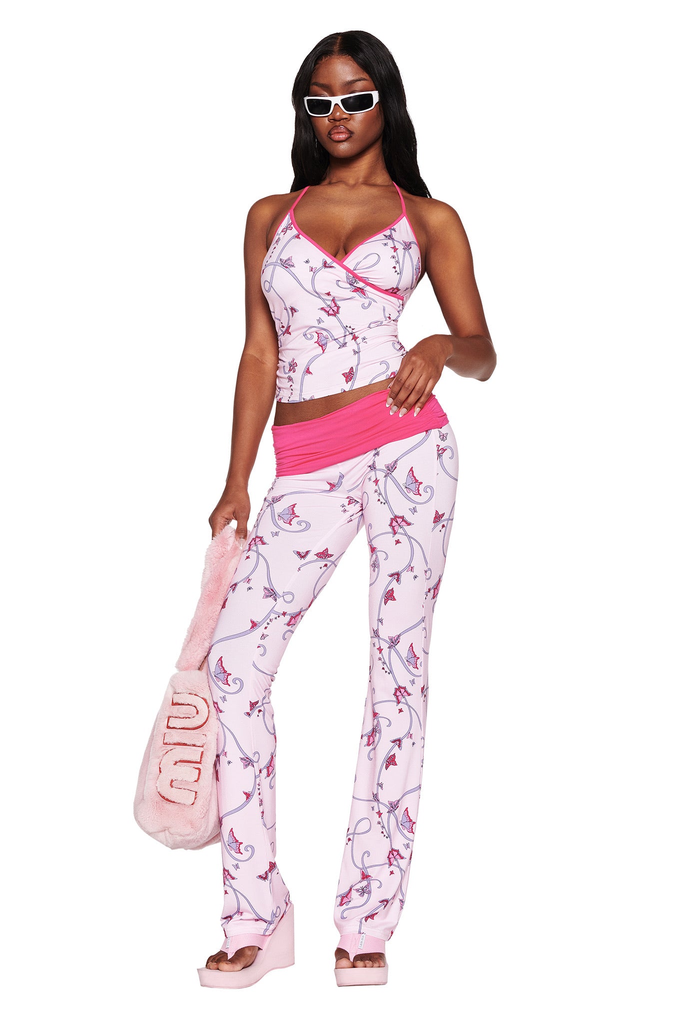 Dressed confidently, the person wears I.AM.GIA's JANNIS TANK in Pink Butterfly Ribbon with matching flared pants and platform sandals, accessorized with stylish sunglasses and a fuzzy pink bag, all crafted from sustainable bamboo.