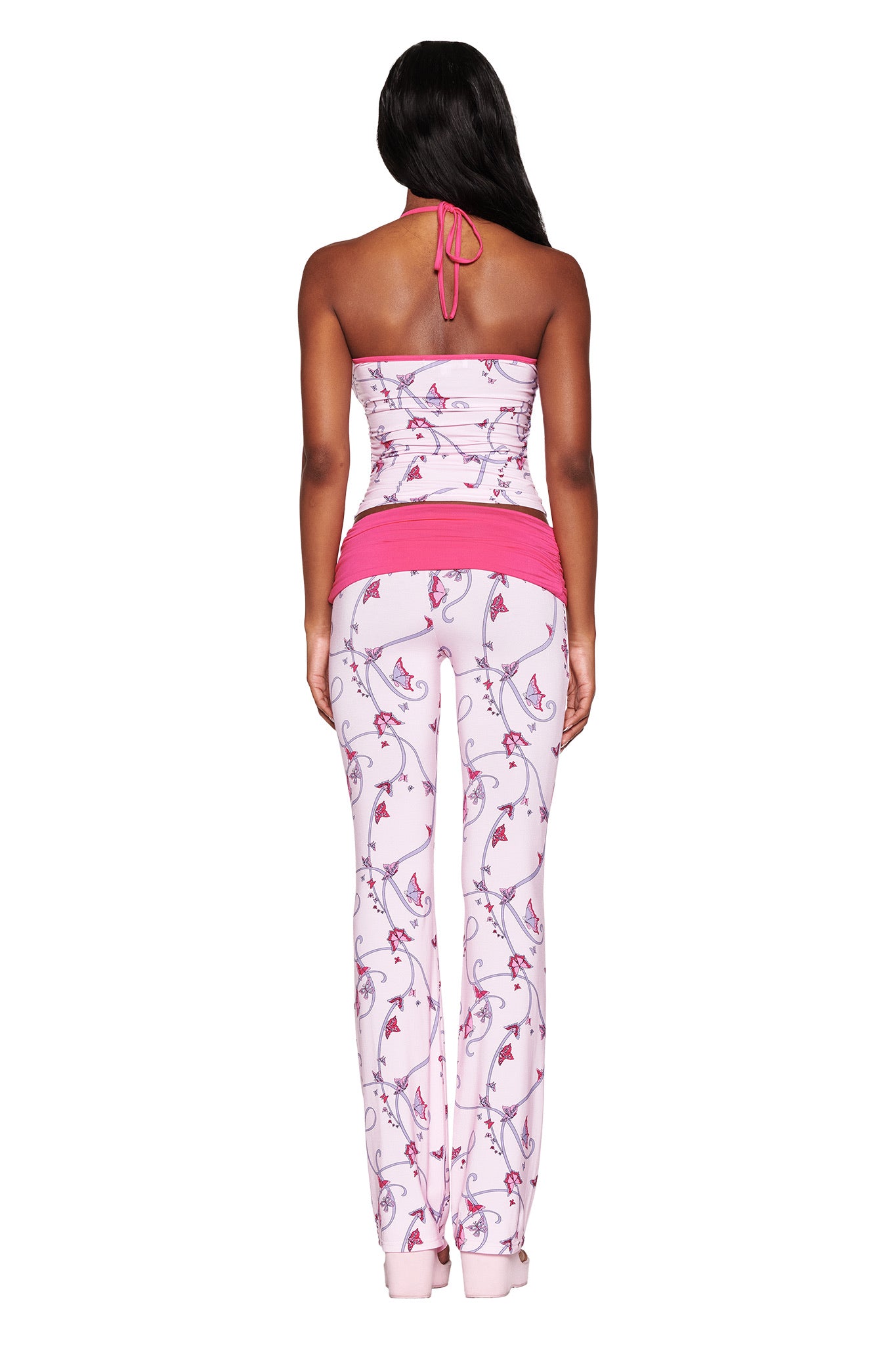 A person stands facing away, wearing I.AM.GIA's Jannis Tank: Pink Butterfly Ribbon, complemented by floral leggings. The strapless bamboo top ties with a halter string at the back, completing the look with a pink waistband and matching ankle trim.