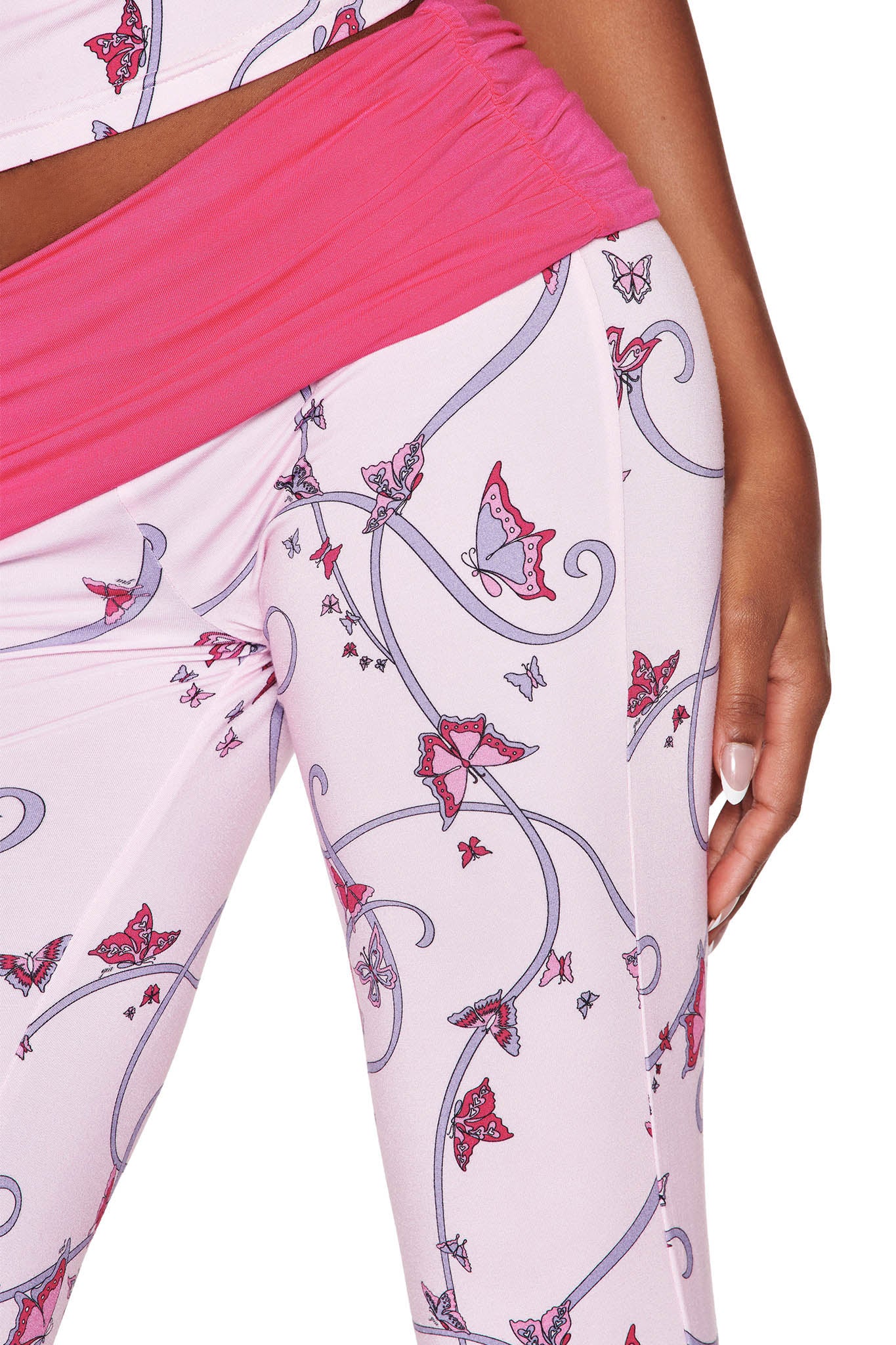 A person is seen wearing the JANNIS PANT - PINK: BUTTERFLY RIBBON by I.AM.GIA, featuring a butterfly ribbon print with a dark pink waistband. The matching top completes their look as their hand rests elegantly by their side.