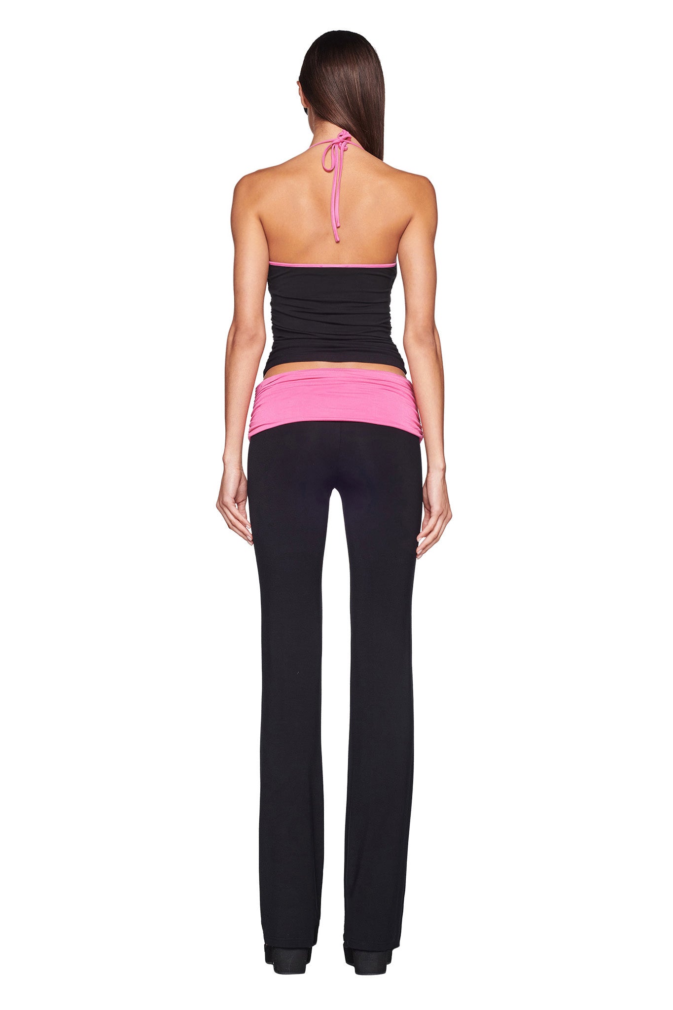 A woman stands facing away, wearing the I.AM.GIA JANNIS PANT in black with a pink waistband and a black halter top. Her long hair falls straight down her back against the plain white background.