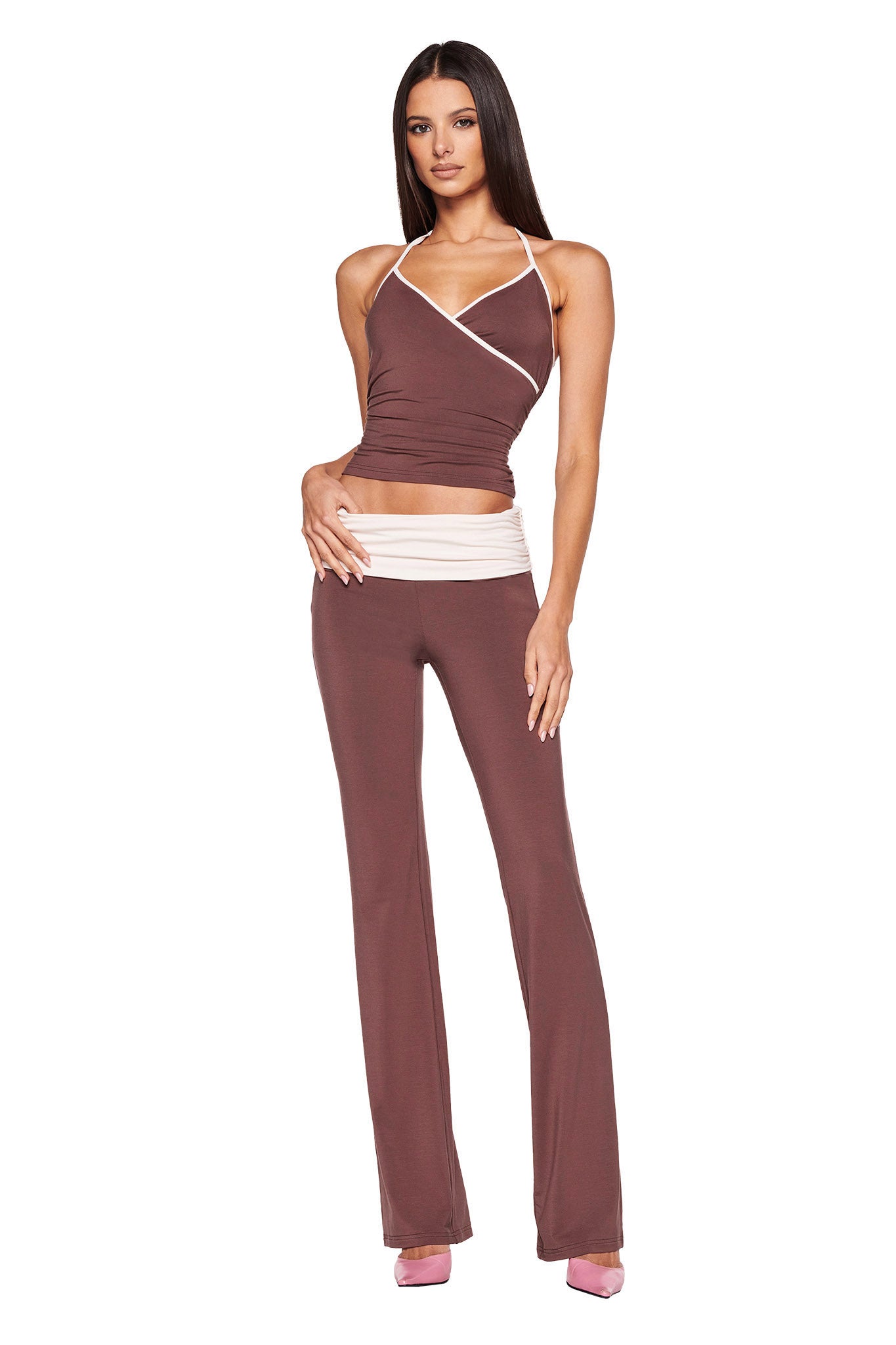 A woman is wearing the JANNIS PANT - BROWN : CHOCOLATE from I.AM.GIA, featuring a rich chocolate hue with a white waistband, matching her brown sleeveless top with white trim. The flared legs of her pants add elegance to her look. She has long, straight hair and stands confidently with one hand on her hip and the other resting by her side, complemented by pink shoes.