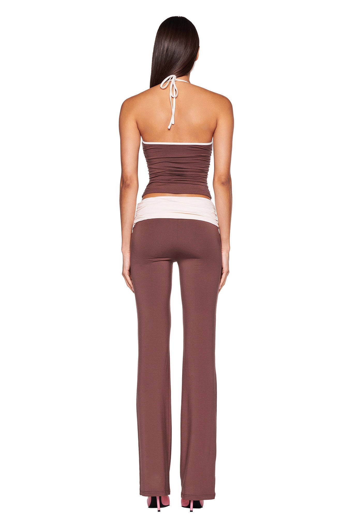 A person with long, dark hair is seen from the back, wearing the JANNIS TANK - BROWN : CHOCOLATE by I.AM.GIA, which features a white halter neck tie. They are also sporting brown flared pants with a white waistband. The bodycon silhouette outfit has a fitted look, and the person stands on a white background.