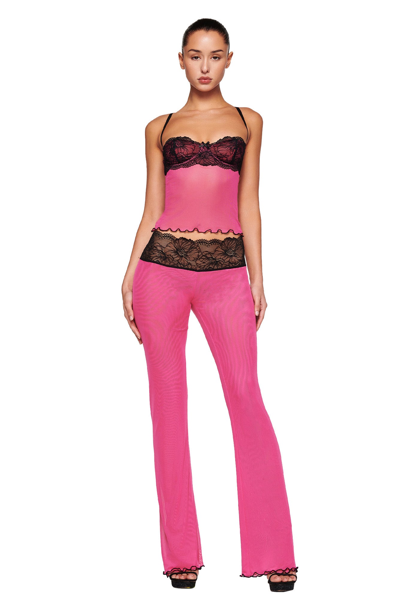 A woman poses confidently in a stylish two-piece outfit from I.AM.GIA. She wears the SILO TOP - PINK, which is adorned with chic black lace detailing around the bust and hem, creating a bustier-like effect. Her ensemble is completed with matching flared mesh pants featuring black lace trim at the waist. Black high heels elevate her look.