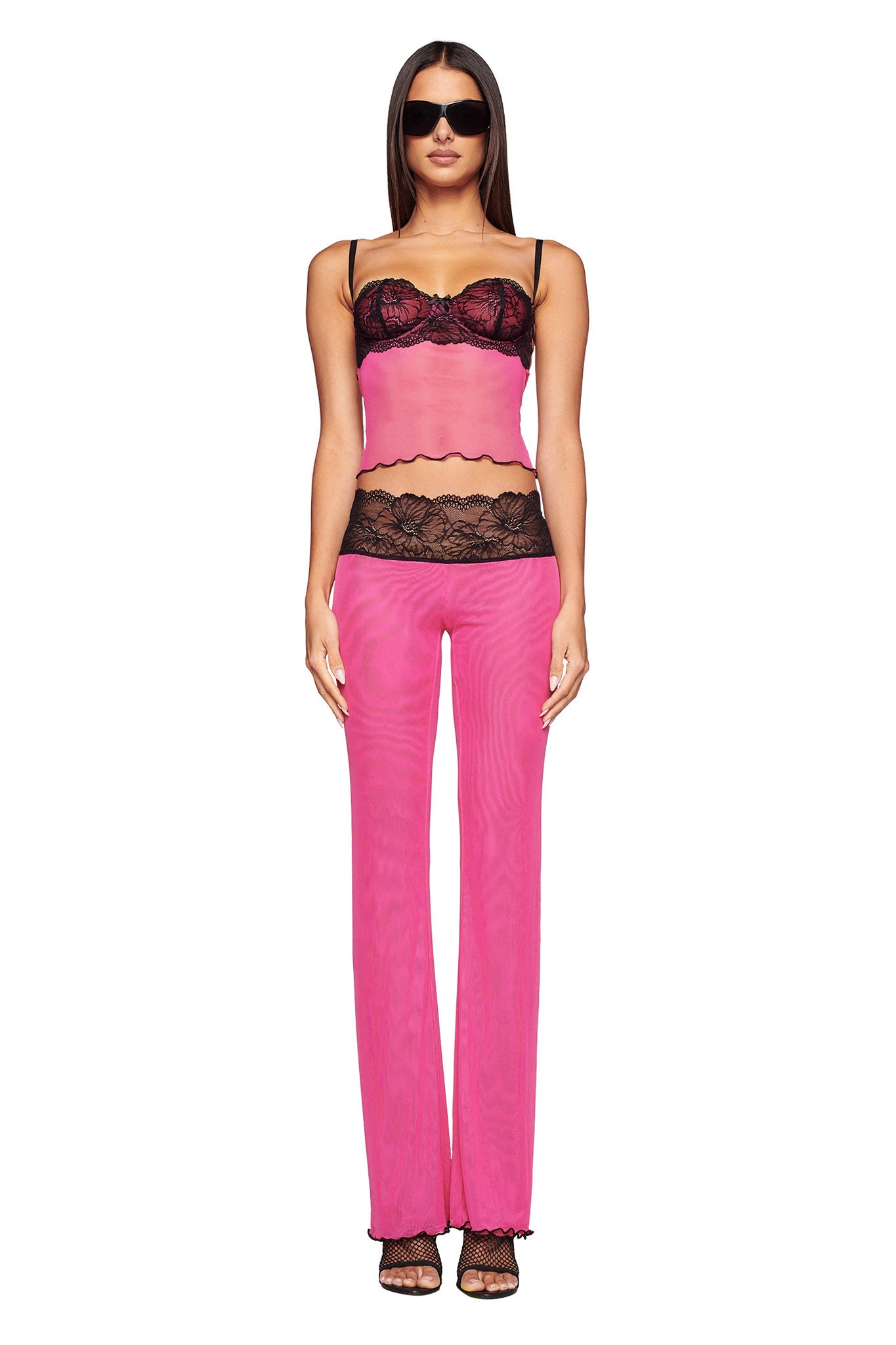 A woman stands against a plain background, showcasing the I.AM.GIA SILO TOP in pink, paired with a matching lingerie set accented with black lace and a black lace bustier. She sports long straight hair, sunglasses, and high-heeled sandals, standing with her hands by her sides.