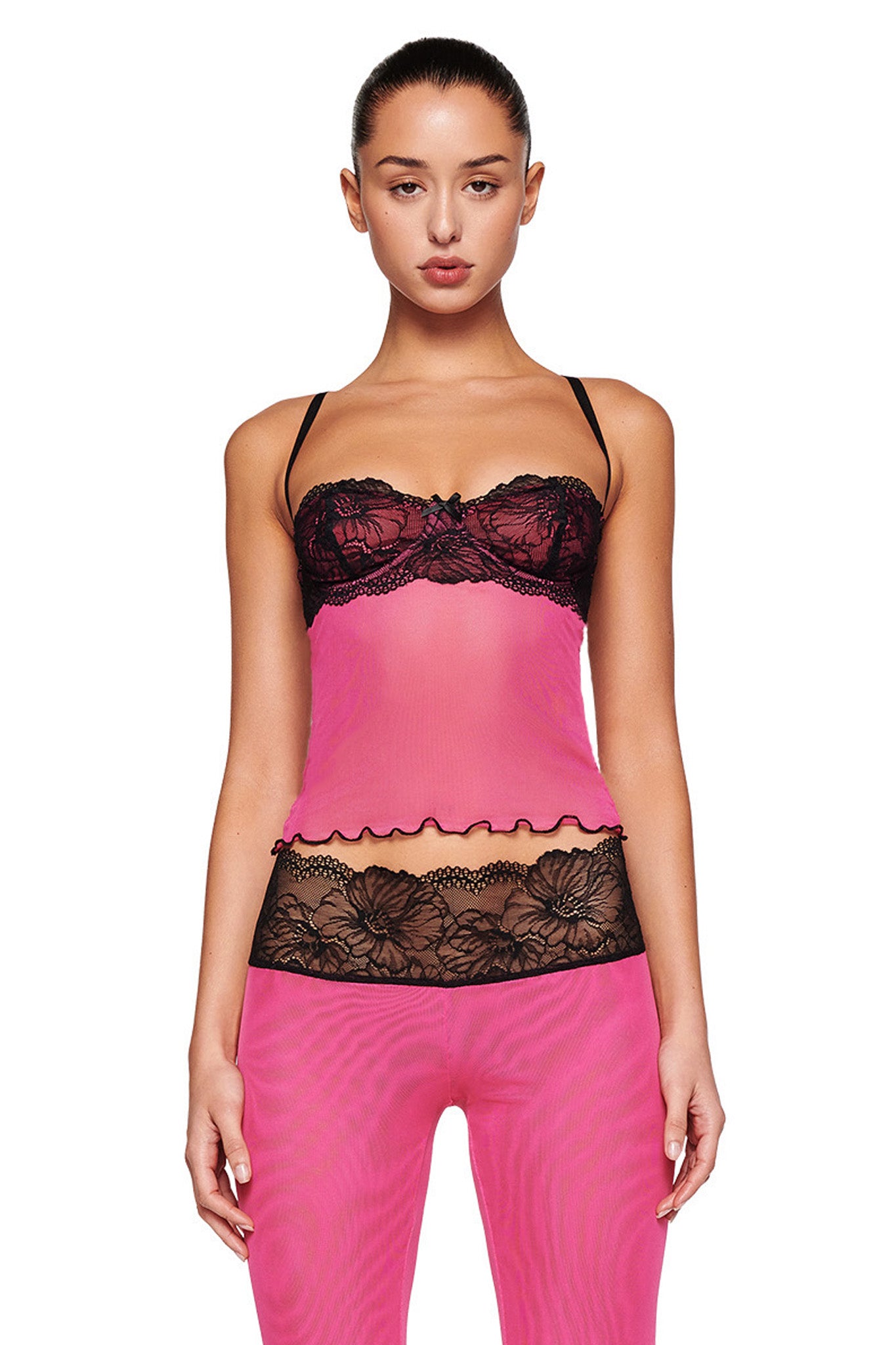 A woman is shown wearing the SILO TOP - PINK by I.AM.GIA. The pink top features black lace detailing over the bustier, with thin black straps and a scalloped hem. She pairs it with a bottom that has a sheer, mesh fabrication waistband in pink. Her hair is styled in a tight bun.