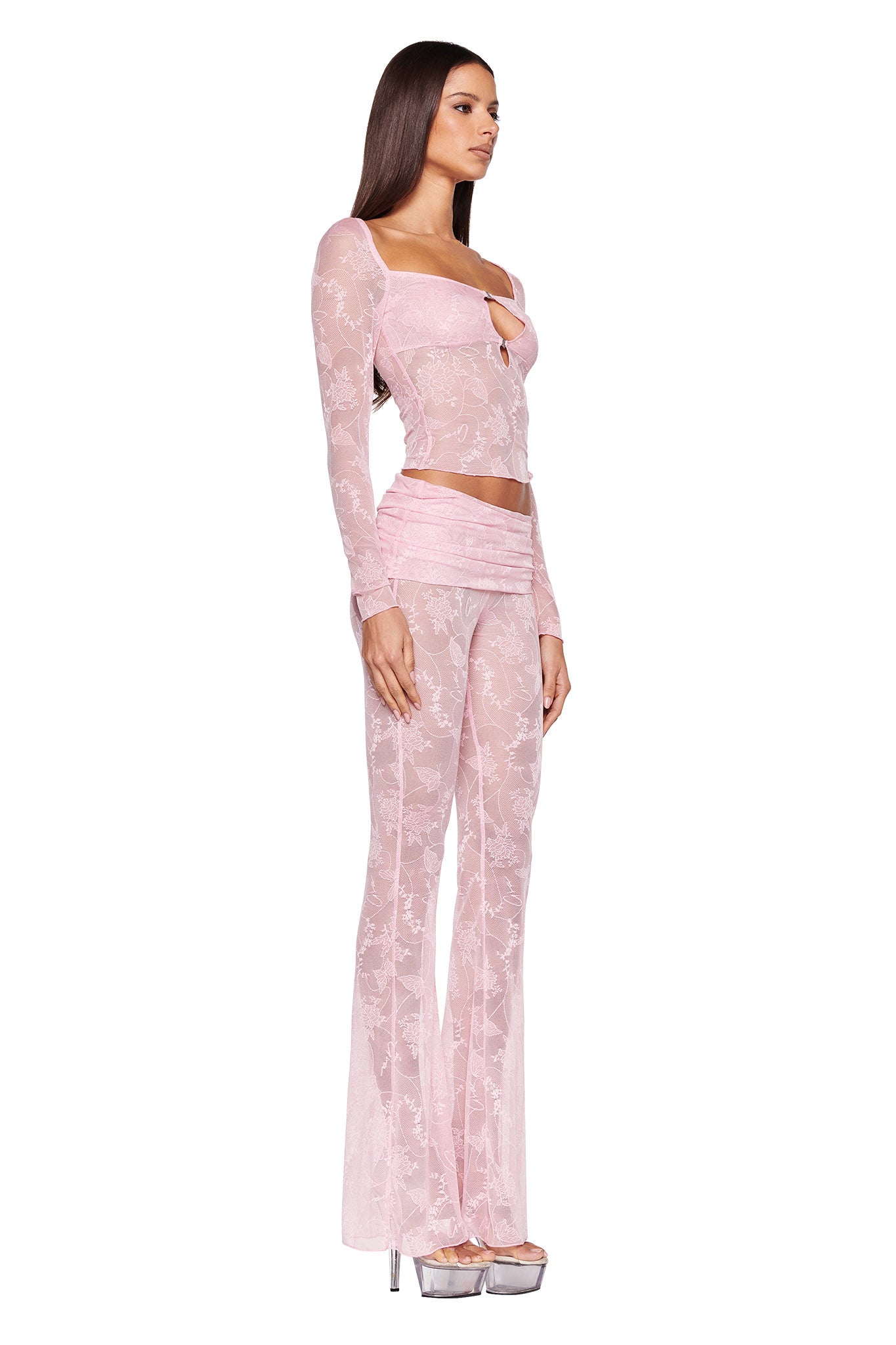 A woman with long brown hair is wearing an I.AM.GIA light pink, sheer, floral-printed long-sleeve top with a keyhole cutout paired with the matching NALLIAH PANT - PINK : LACE. She is standing in profile, facing to the right, and is wearing silver high-heeled platform shoes.
