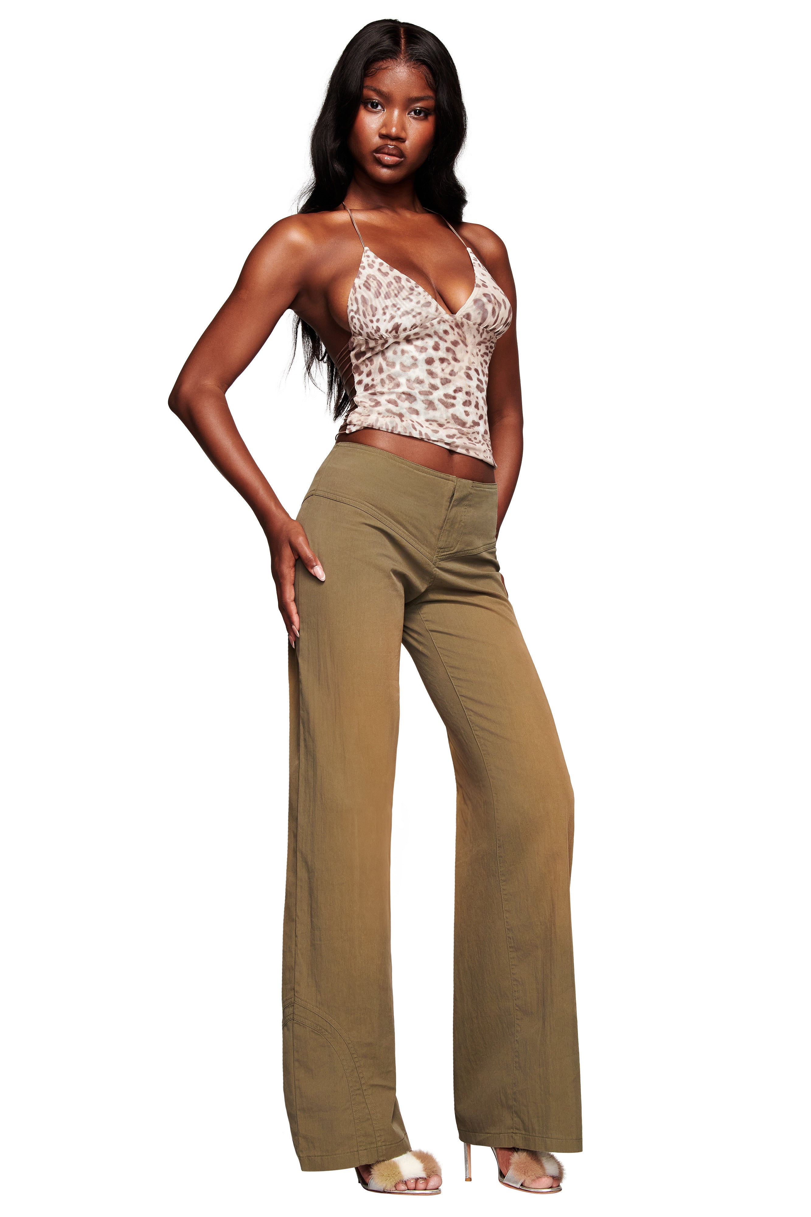A woman stands confidently in the I.AM.GIA ELLERY TOP - LEOPARD, featuring a sleeveless mesh halter design, paired with high-waisted wide-leg olive green pants and beige high heels. She poses with one hand on her hip against a white background.