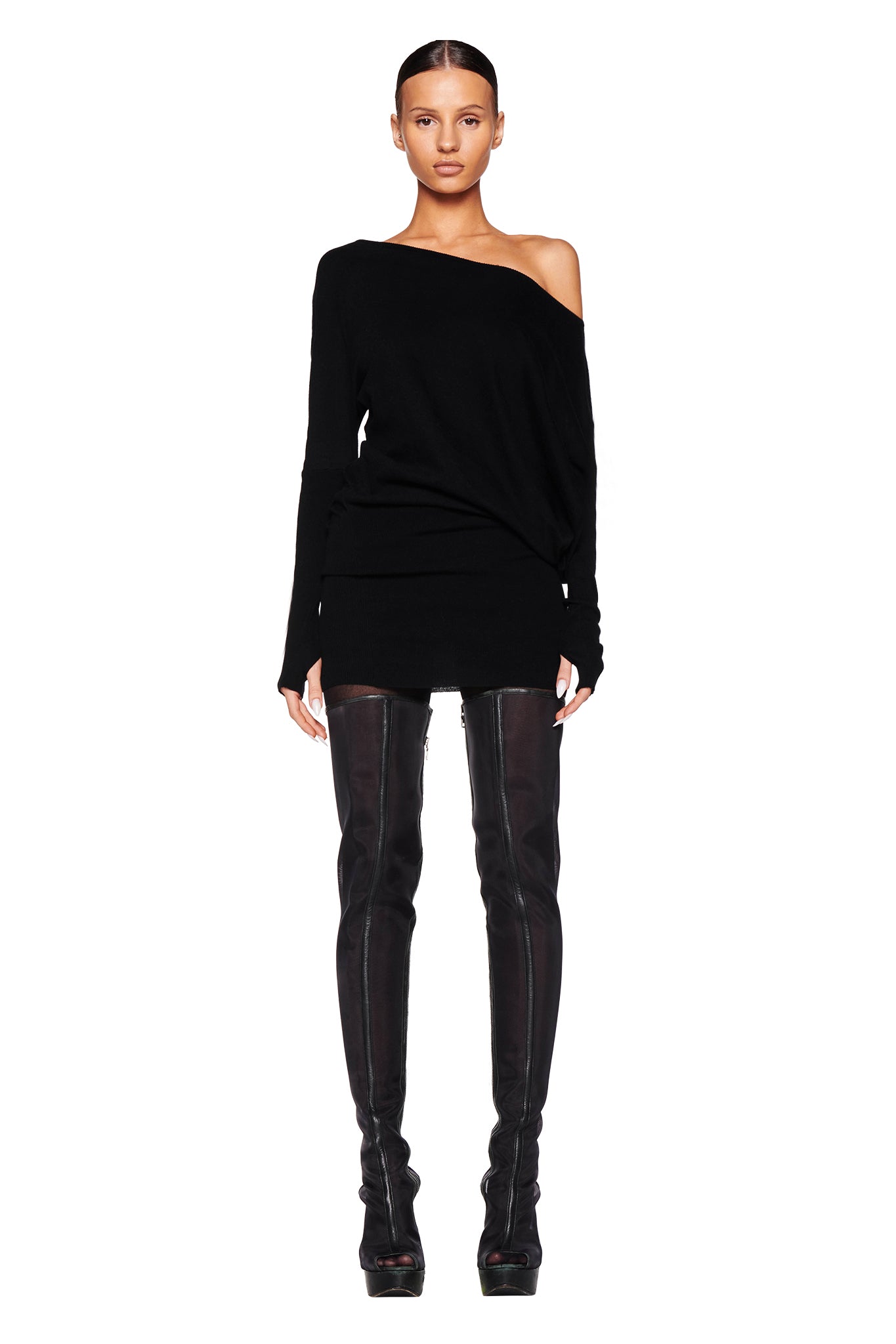 A woman stands against a white background wearing the GISELLE MINI DRESS - BLACK from I.AM.GIA, which features an asymmetrical design that exposes one shoulder. She pairs the dress with thigh-high black boots, her hair pulled back, and presents a neutral expression.