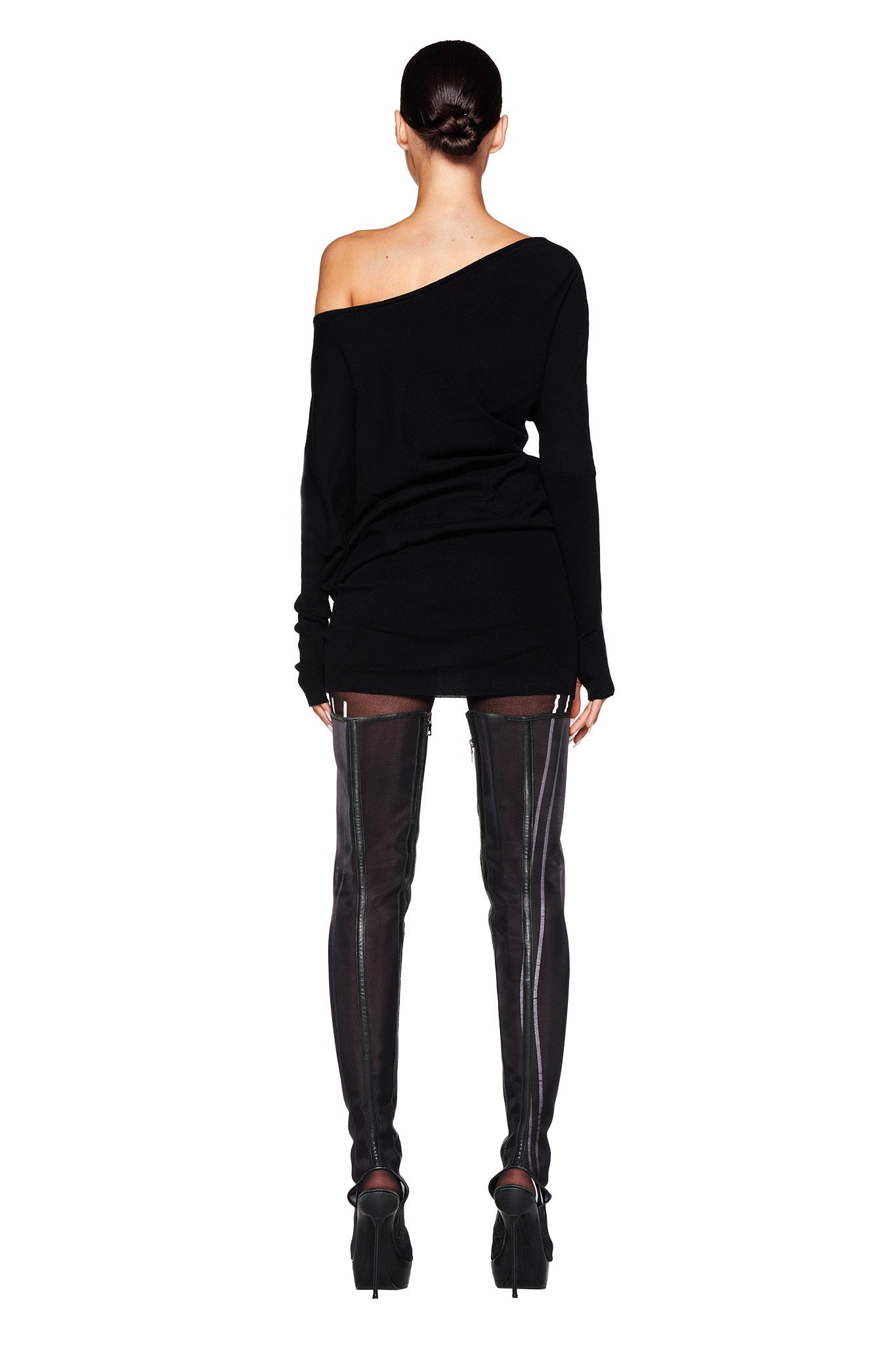 A person stands with their back to the camera, wearing the GISELLE MINI DRESS in black from I.AM.GIA and thigh-high black boots with visible front zippers. The individual's hair is pulled back into a sleek bun against a white background.