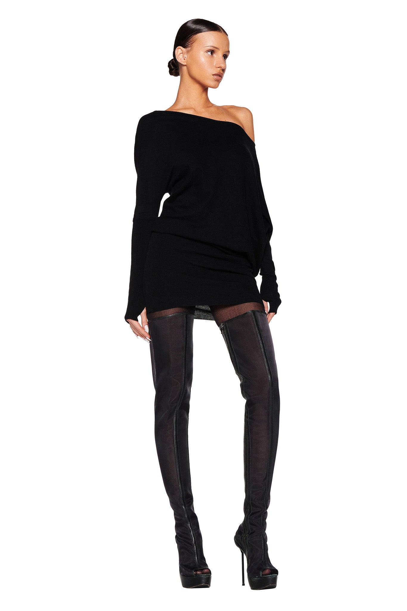 A model stands against a plain white background, wearing the GISELLE MINI DRESS in black from I.AM.GIA and black thigh-high boots with high heels. Her hair is slicked back, and she poses with her right leg slightly forward, and her right hand resting on her hip.