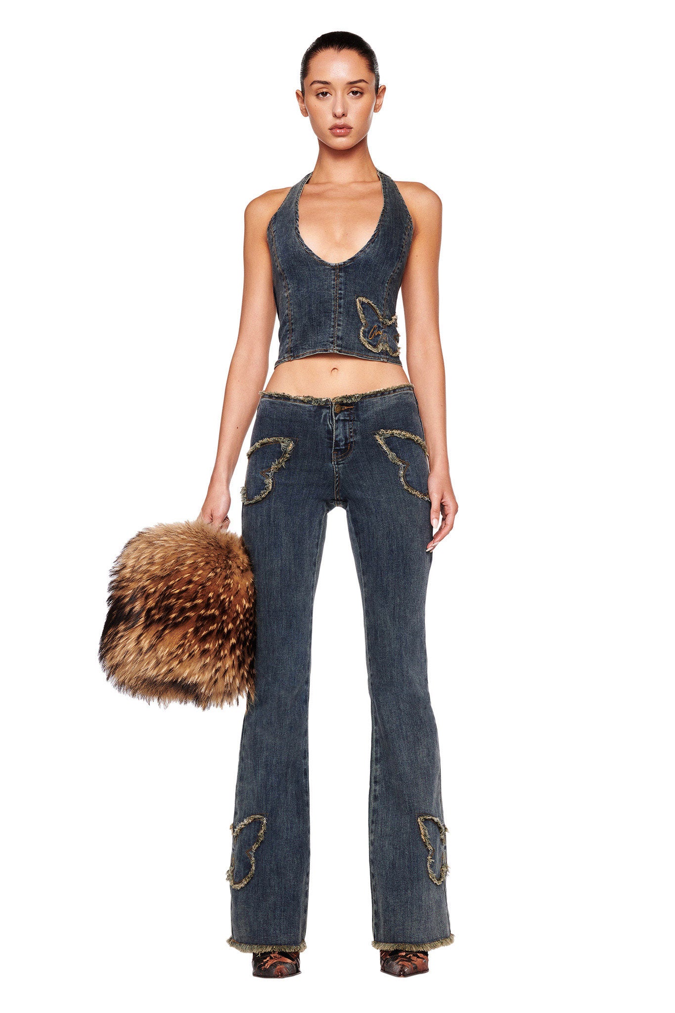 A woman stands facing forward wearing a halter-style stretch denim top paired with the I.AM.GIA SAFFIYAH PANT in denim blue, featuring low-rise waist bell-bottoms adorned with butterfly appliques. She holds a fluffy brown and black furry bag in her left hand. Her sleek hair is tied back, and she stands against a white background.