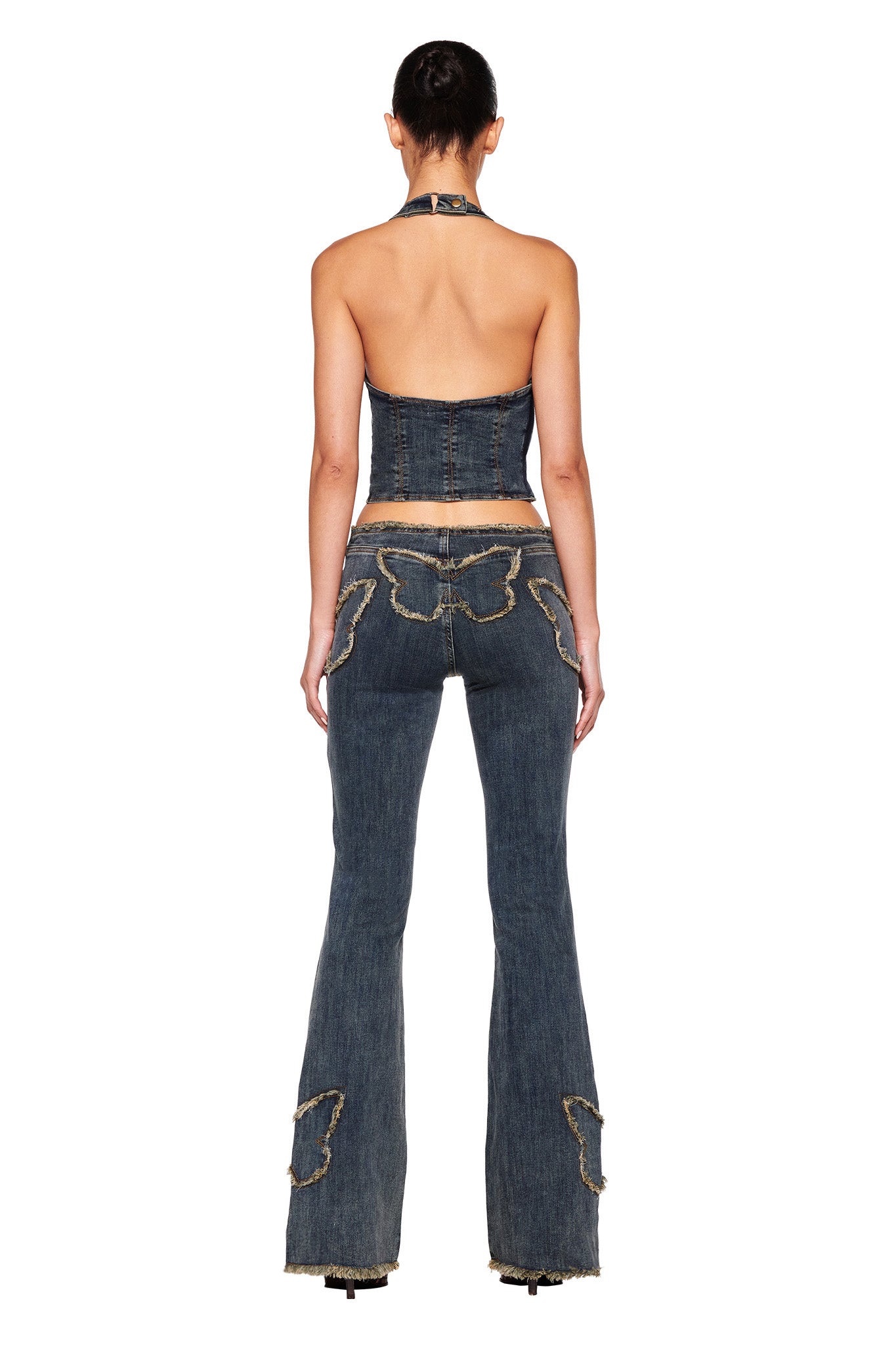 A woman faces away from the camera, dressed in a dark denim halter top paired with the I.AM.GIA SAFFIYAH PANT - BLUE. The denim blue, low-rise waist jeans feature butterfly designs stitched on the back pockets and lower legs. Her hair is styled in a bun, and she stands against a plain white background.